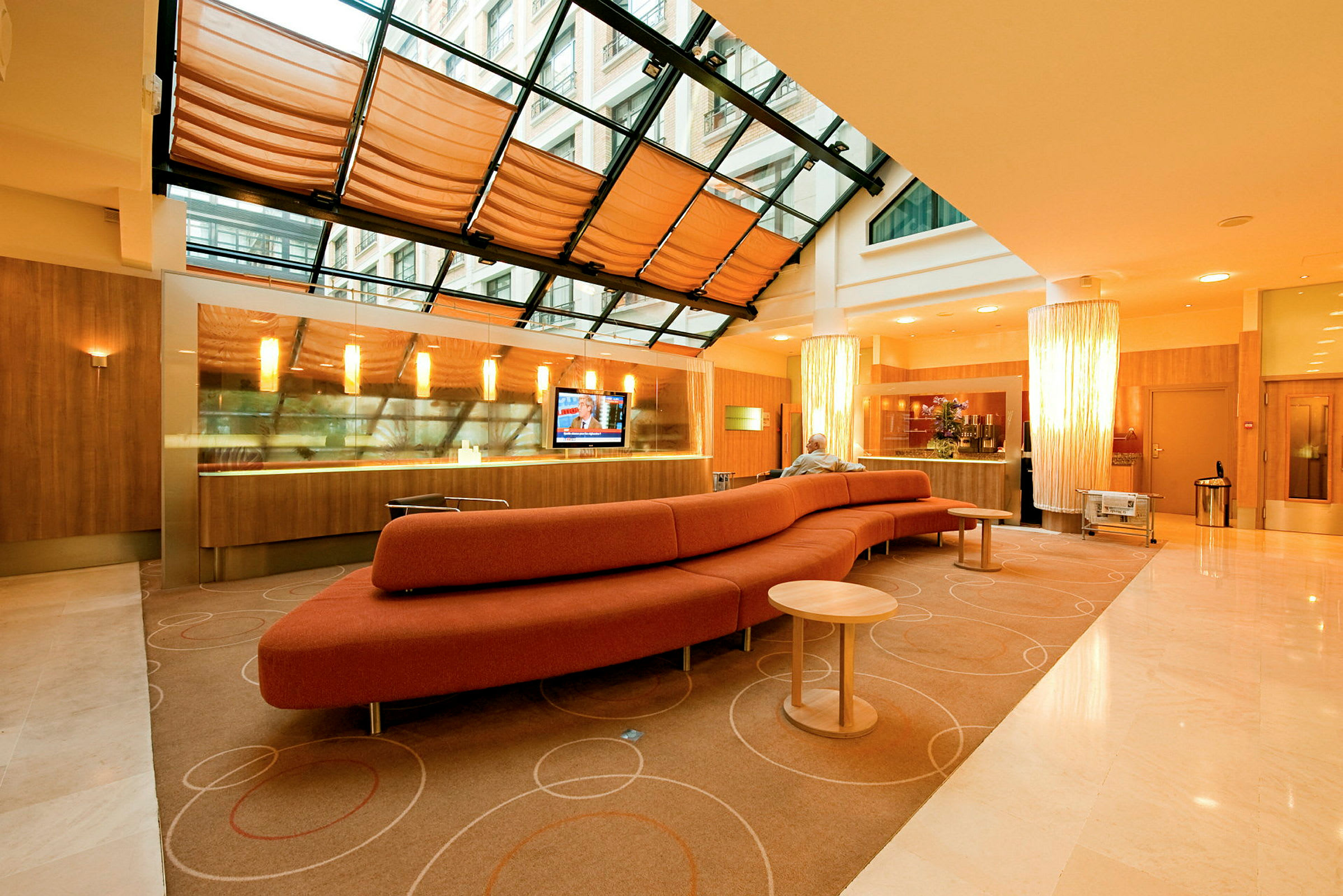 Et moderne hotellobbyområde med en lang rød sofa, et glasstak med gardiner og et resepsjonsområde i bakgrunnen. (AI-generert tekst)
