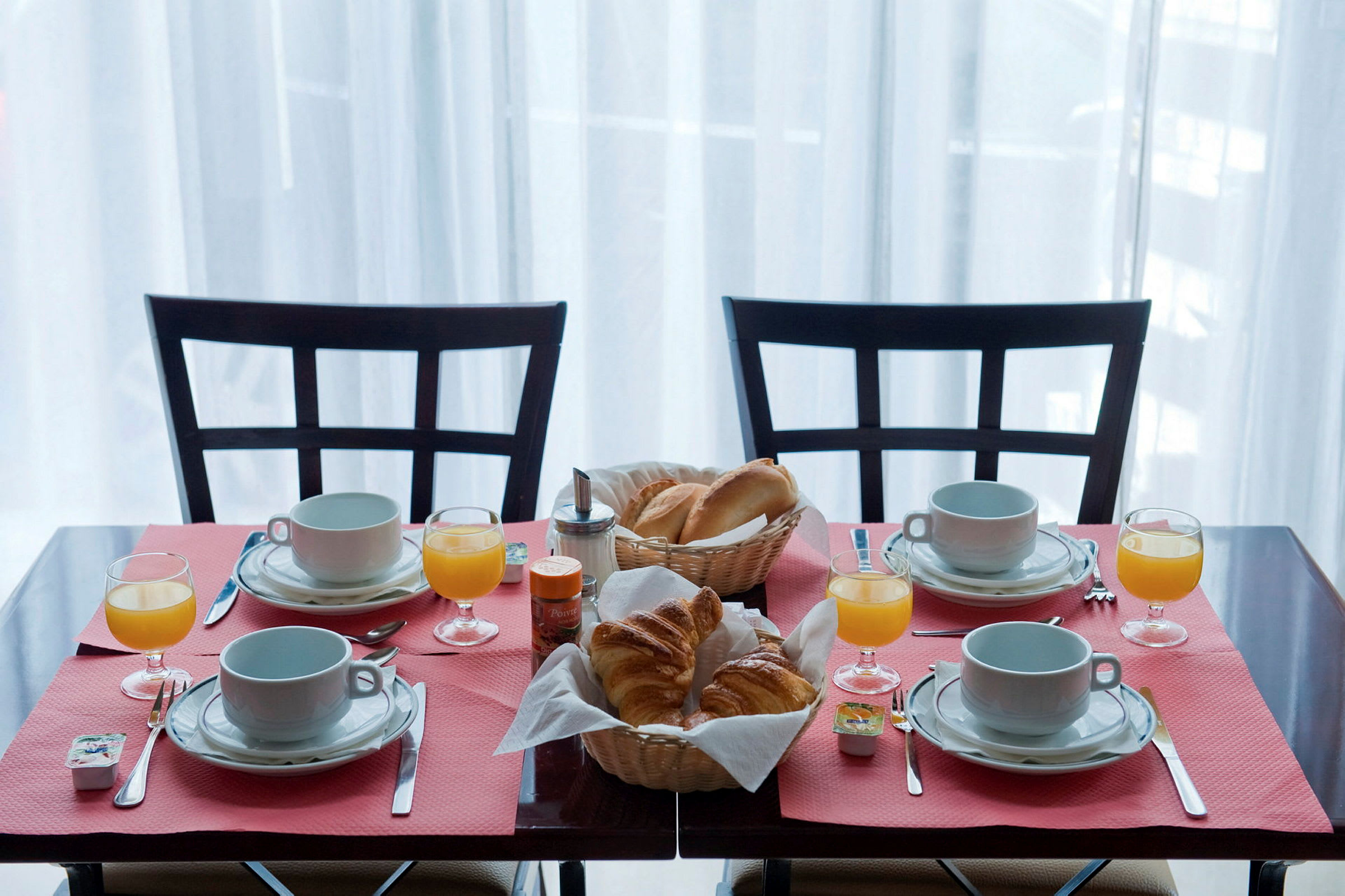 Et frokostbord dekket for fire personer med kaffekopper, glass med appelsinjuice og kurver med brød og croissanter. (AI-generert tekst)