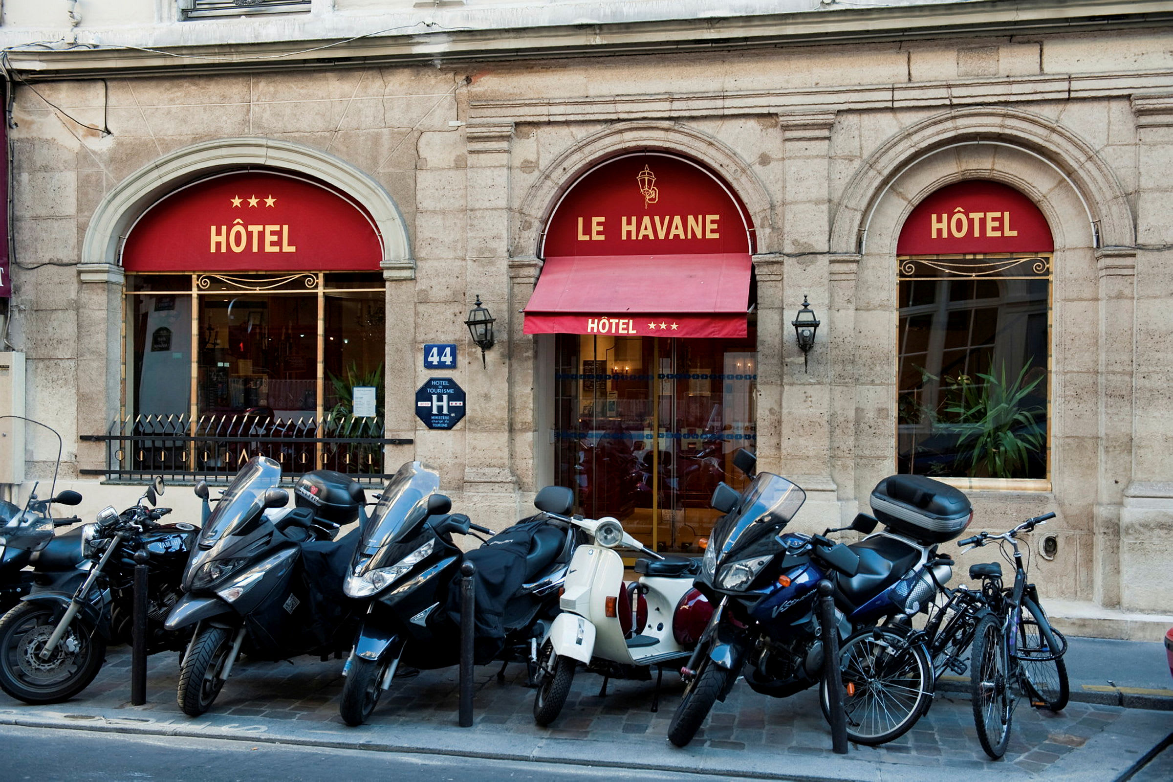 Bilde som viser inngangen til et hotell med navnet Le Havane, tre stjerner vises på markisen. Foran bygningen står mopeder og sykler parkert. (AI-generert tekst)