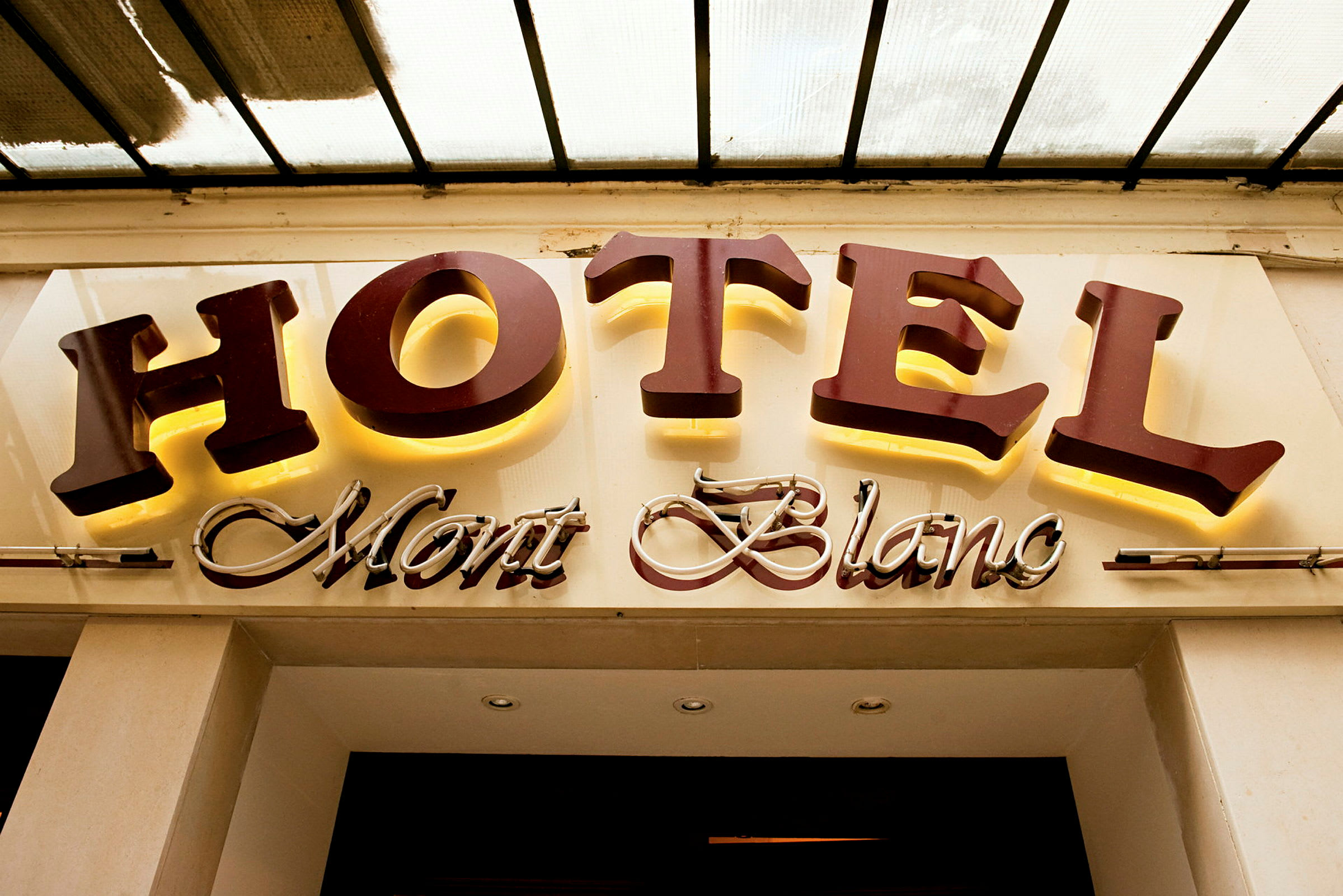 Et hotellskilt med teksten "HOTEL Mont Blanc", der "HOTEL" er i store opplyste bokstaver og "Mont Blanc" er i en kursiv dekorativ stil. (AI-generert tekst)