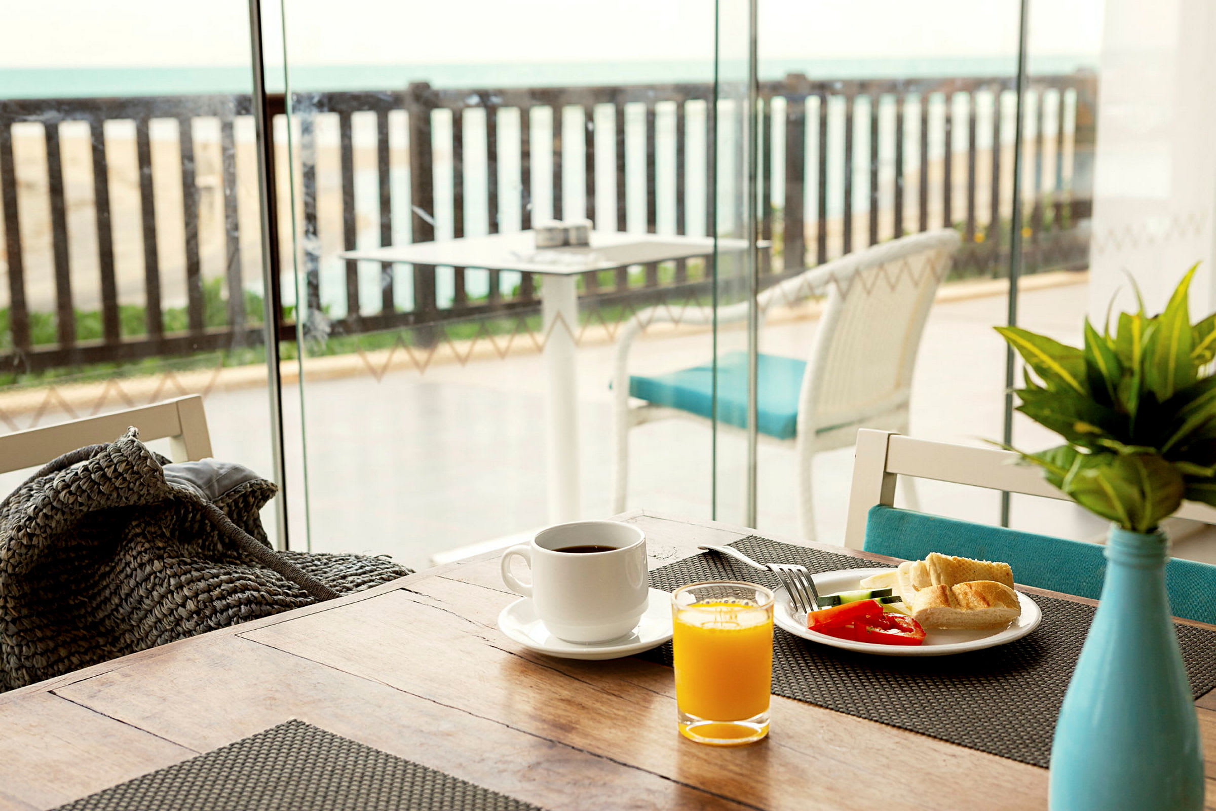 Frokostbord med kaffe, juice og grønnsaker nær et glassvindu med utsikt over en terrasse med havet i bakgrunnen. (AI-generert tekst)
