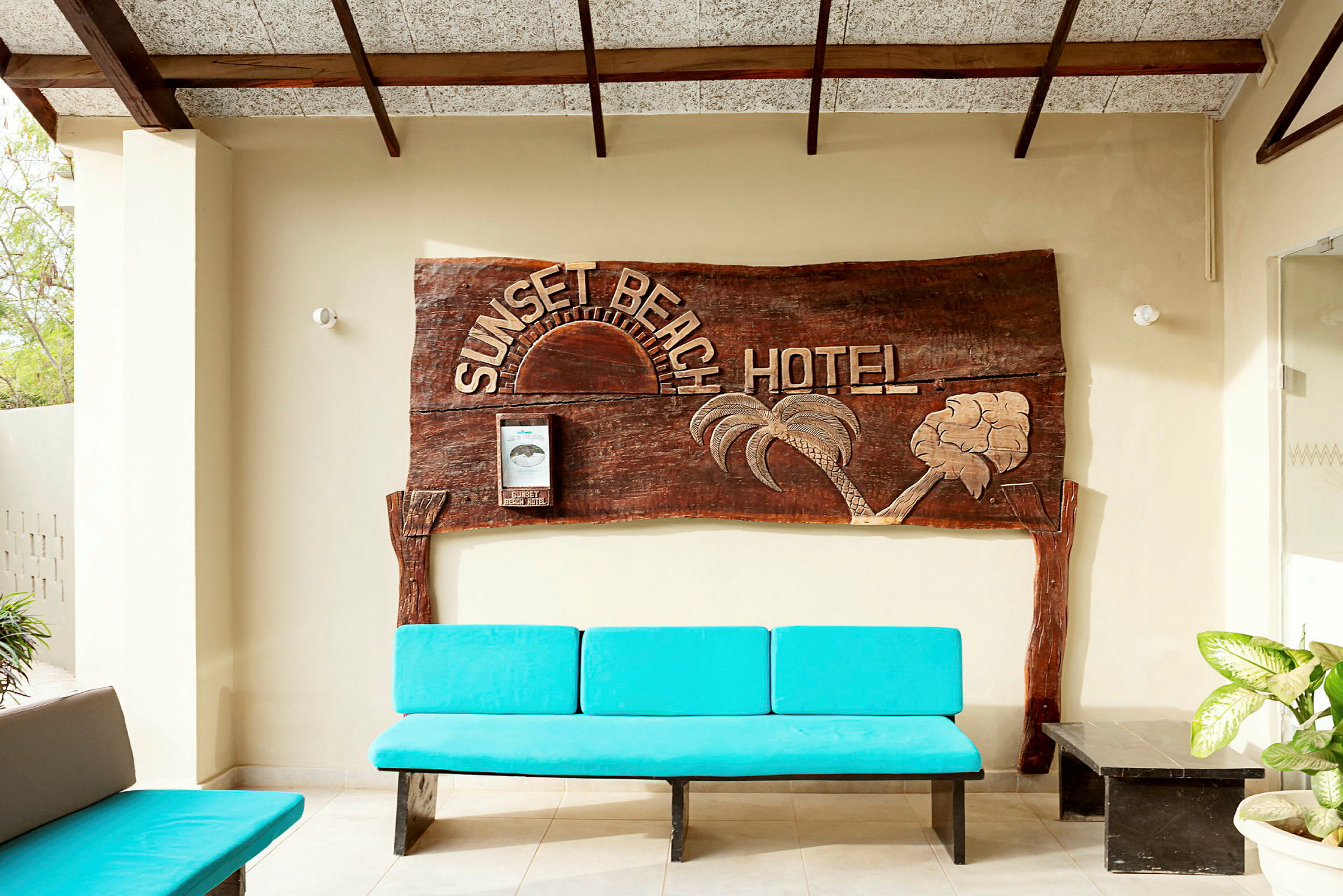 Et treskilt med teksten "Sunset Beach Hotel" pryder veggen. Foran er det en blå benk og noen planter. (AI-generert tekst)