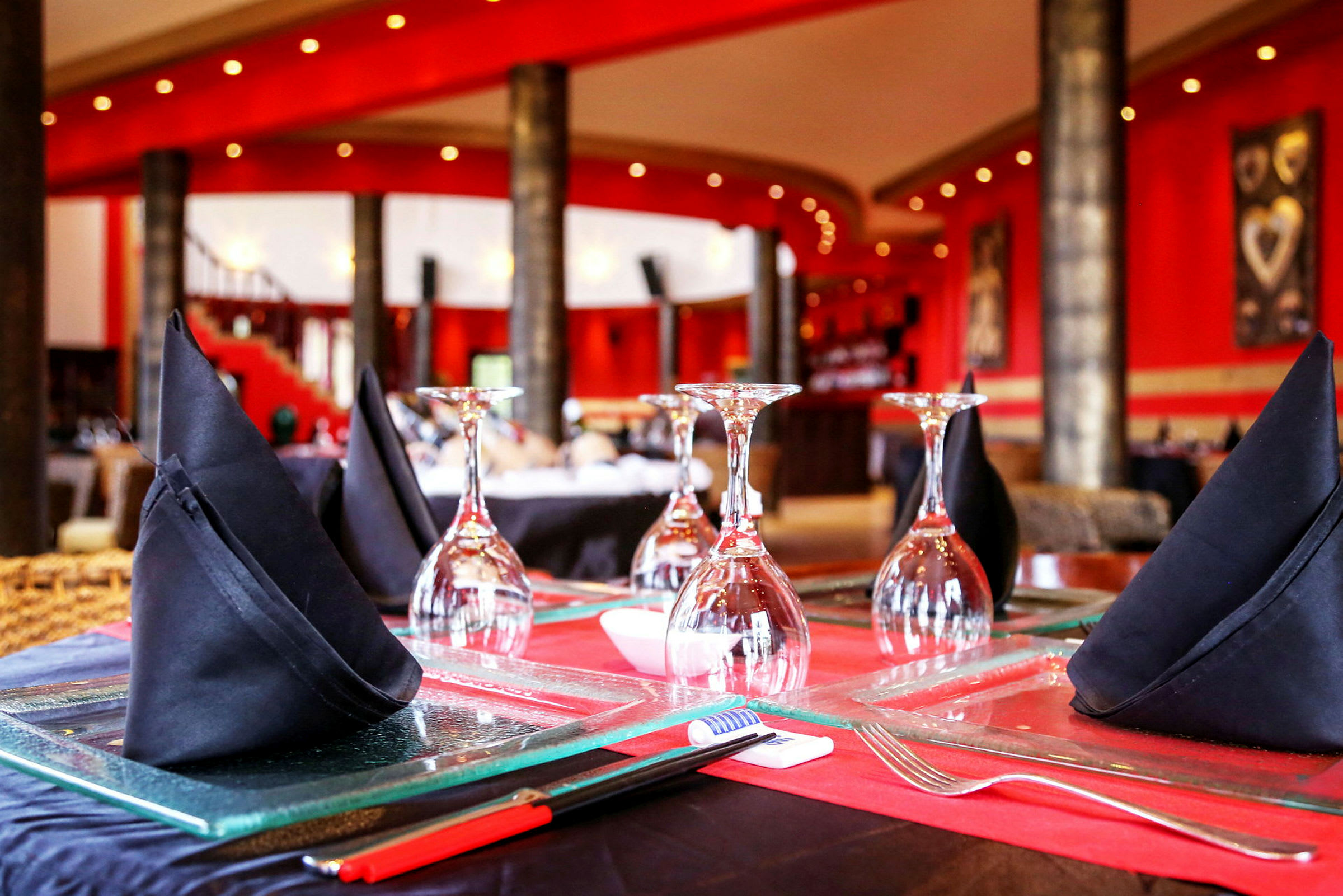 Nærbilde av en restaurantoppsett med glass og svarte stoffservietter på et bord, omgitt av stilfulle røde vegger. (AI-generert tekst)