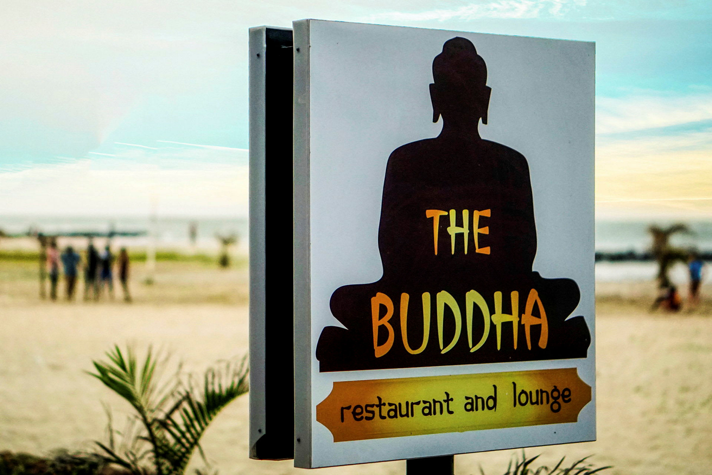 Et skilt med teksten 'The Buddha restaurant and lounge' med en silhuett av en sittende Buddha foran en strandbakgrunn. (AI-generert tekst)