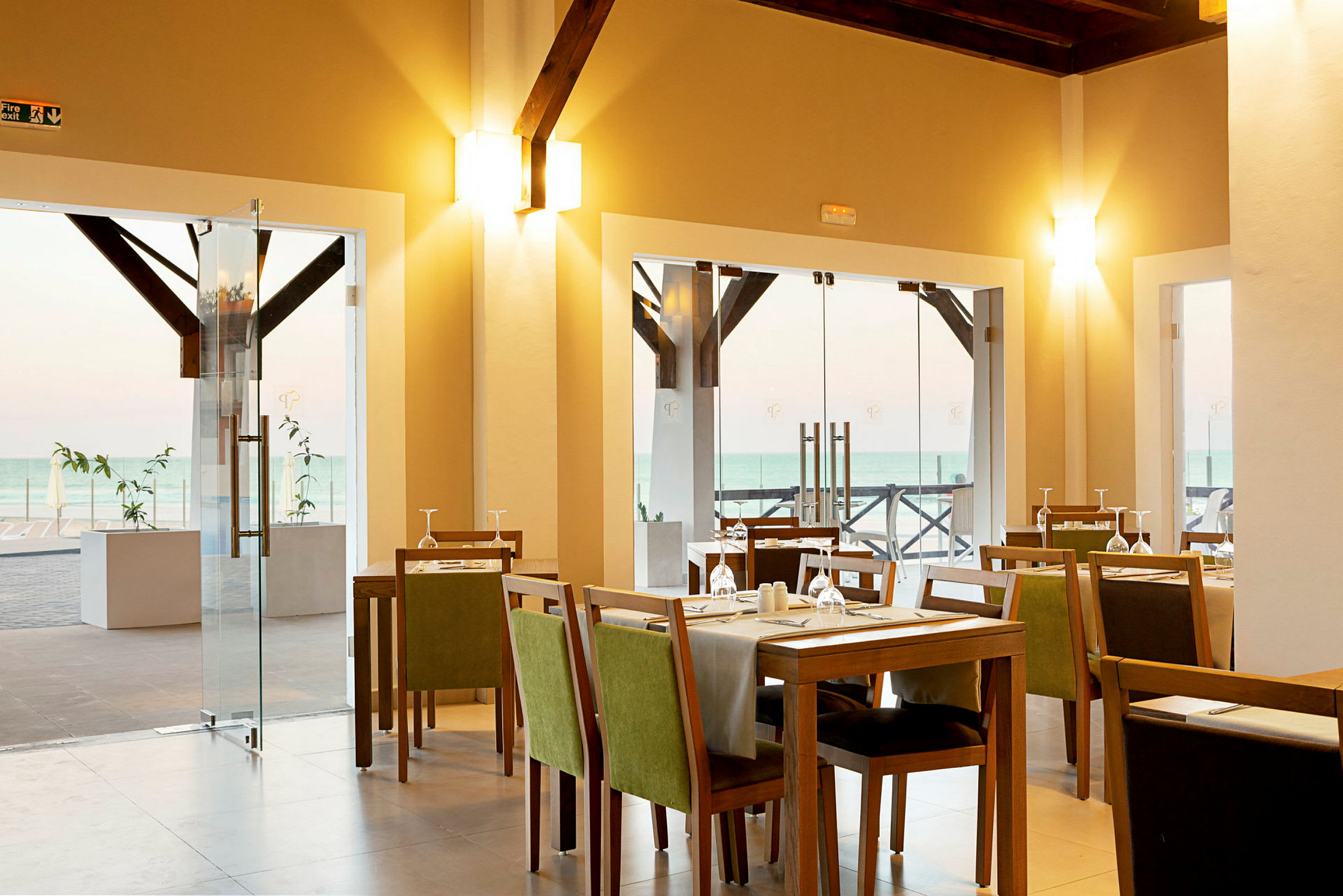 Interiør av en restaurant med bord, stoler og duker, samt glassdører med havutsikt utenfor. (AI-generert tekst)