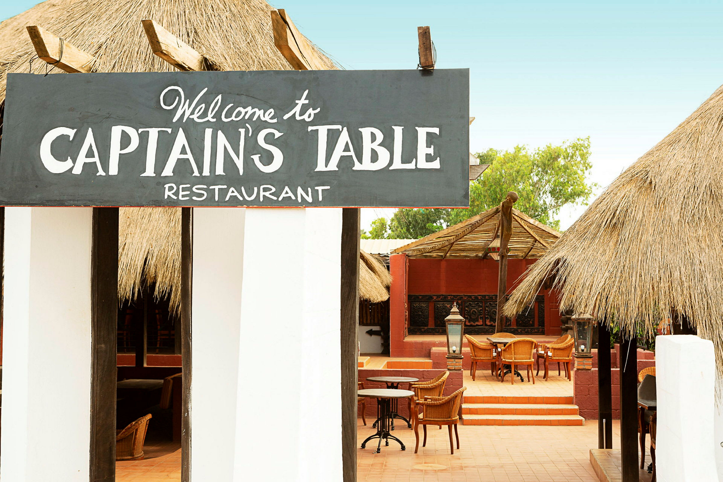 Et restaurantområde med skiltet "Welcome to Captain's Table Restaurant", stoler og bord synlige utendørs under tak. (AI-generert tekst)