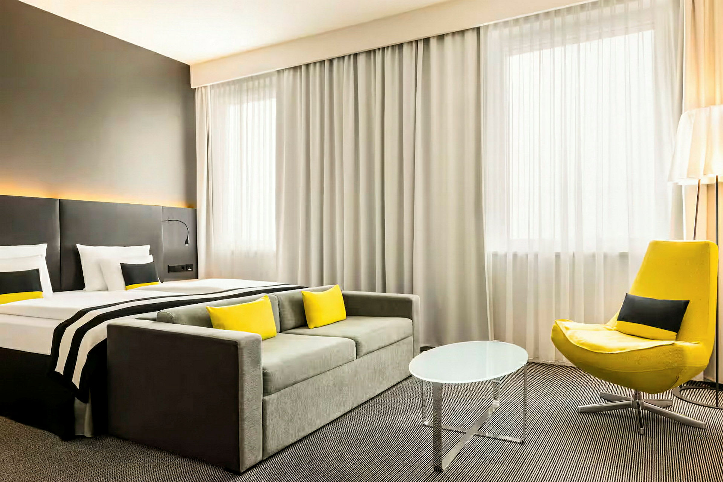 Et hotellrom med en seng, sofa, gul lenestol og et lite rundt bord. (AI-generert tekst)