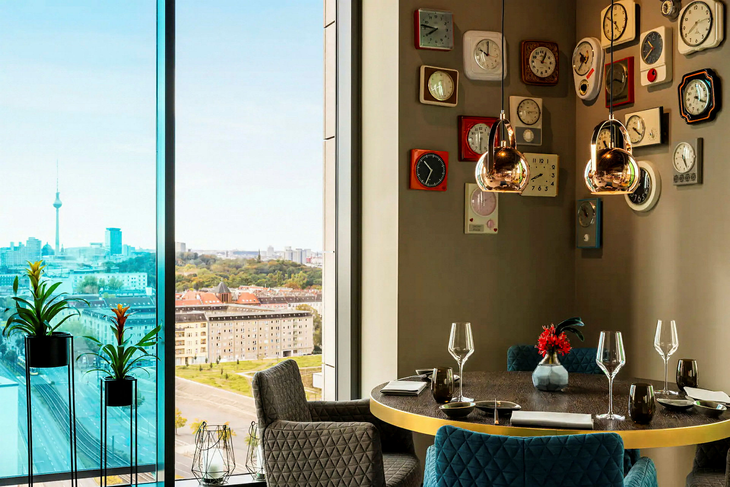 Et moderne rom med store vinduer som viser en urban utsikt med et tårn, omgitt av høye bygninger og grøntområder. Interiøret inkluderer et rundt spisebord med glass og servise samt en vegg dekorert med retro-inspirerte klokker. (AI-generert tekst)