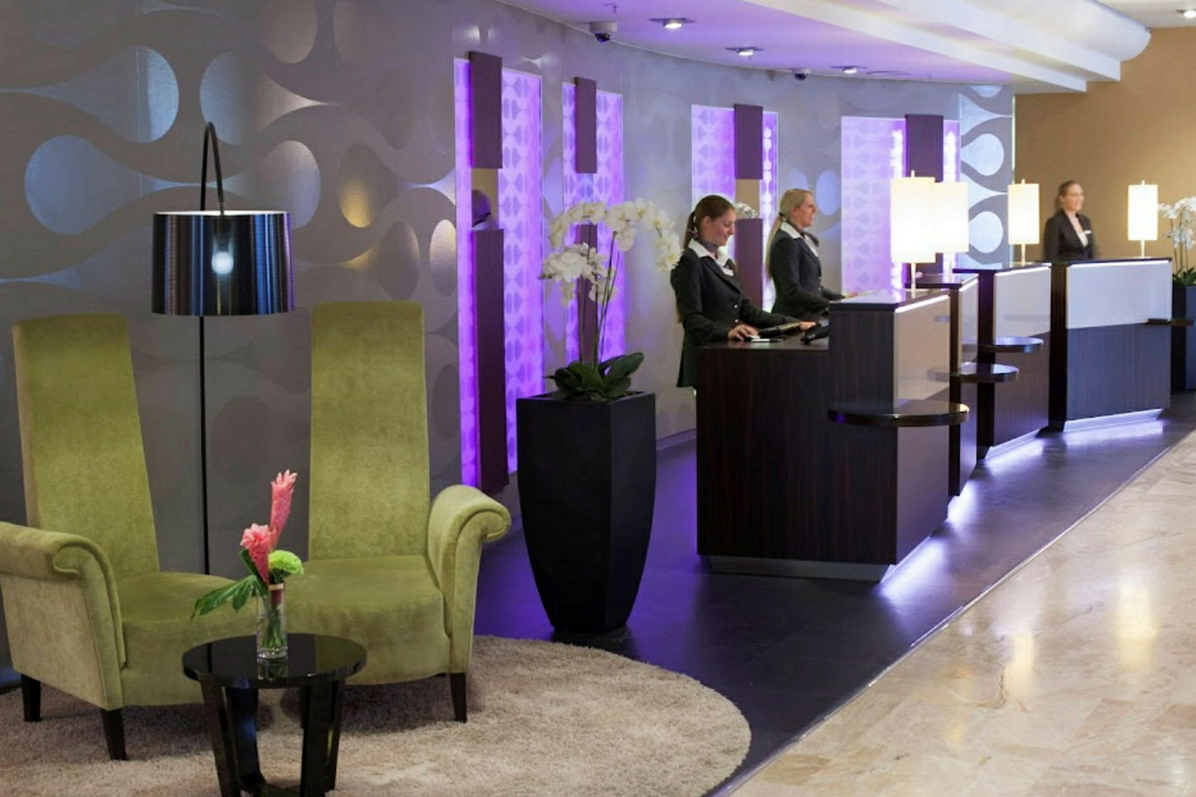 En hotellobby med to grønne stoler, et lite bord med en blomst, og resepsjonsdisker med belysning. (AI-generert tekst)