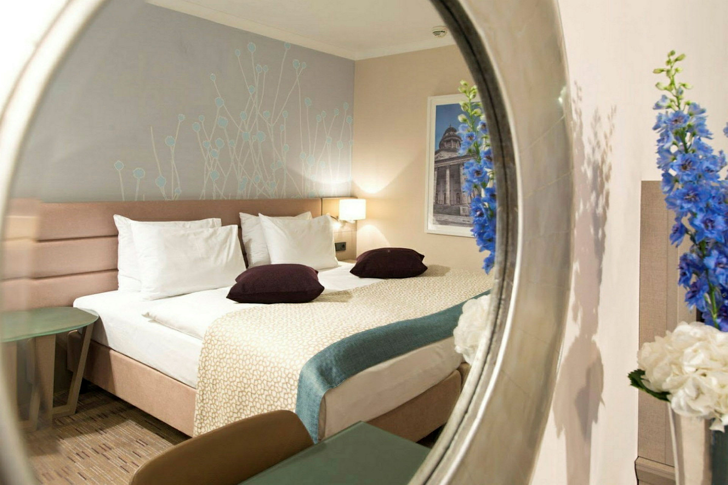 Hotellrom med seng vist gjennom et speil, dekorert med blomster og en veggmaleri. (AI-generert tekst)