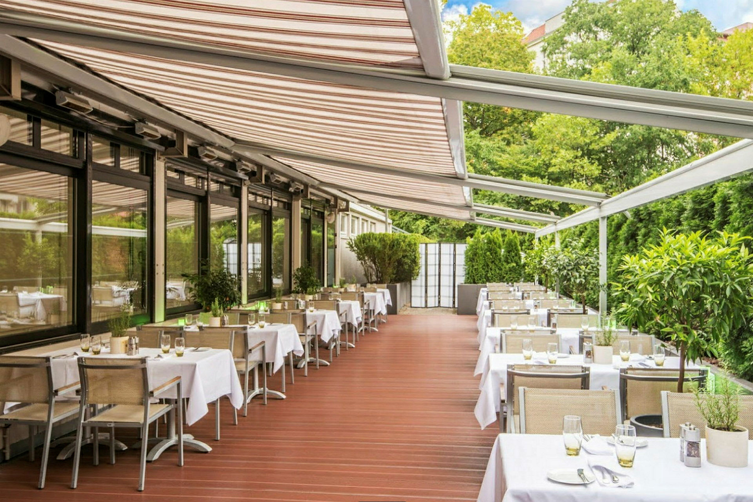 Bilde av en restaurant med bord og stoler under en pergola med grønne planter rundt seg. (AI-generert tekst)
