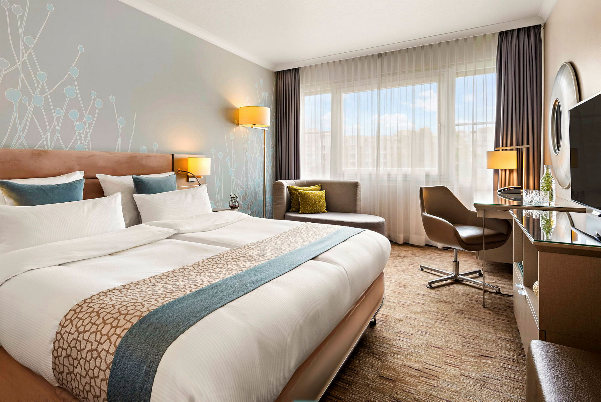Et moderne hotellrom med en stor dobbeltseng, skrivebord, stol og en sittegruppe ved et vindu. (AI-generert tekst)