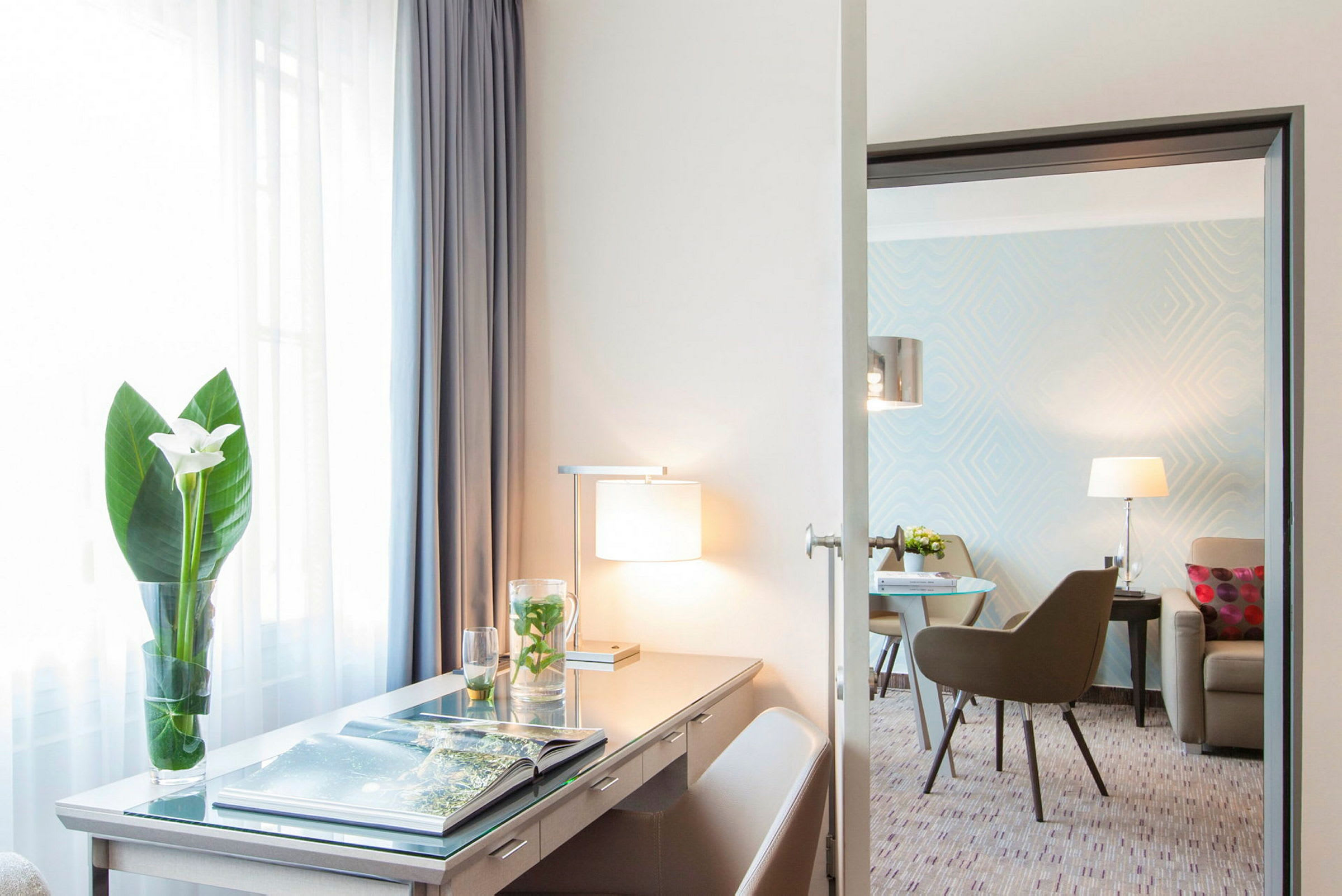 Et hotellrom med et skrivebord, lampe og en blomsterkrukke, samt en sittegruppe i bakgrunnen. (AI-generert tekst)