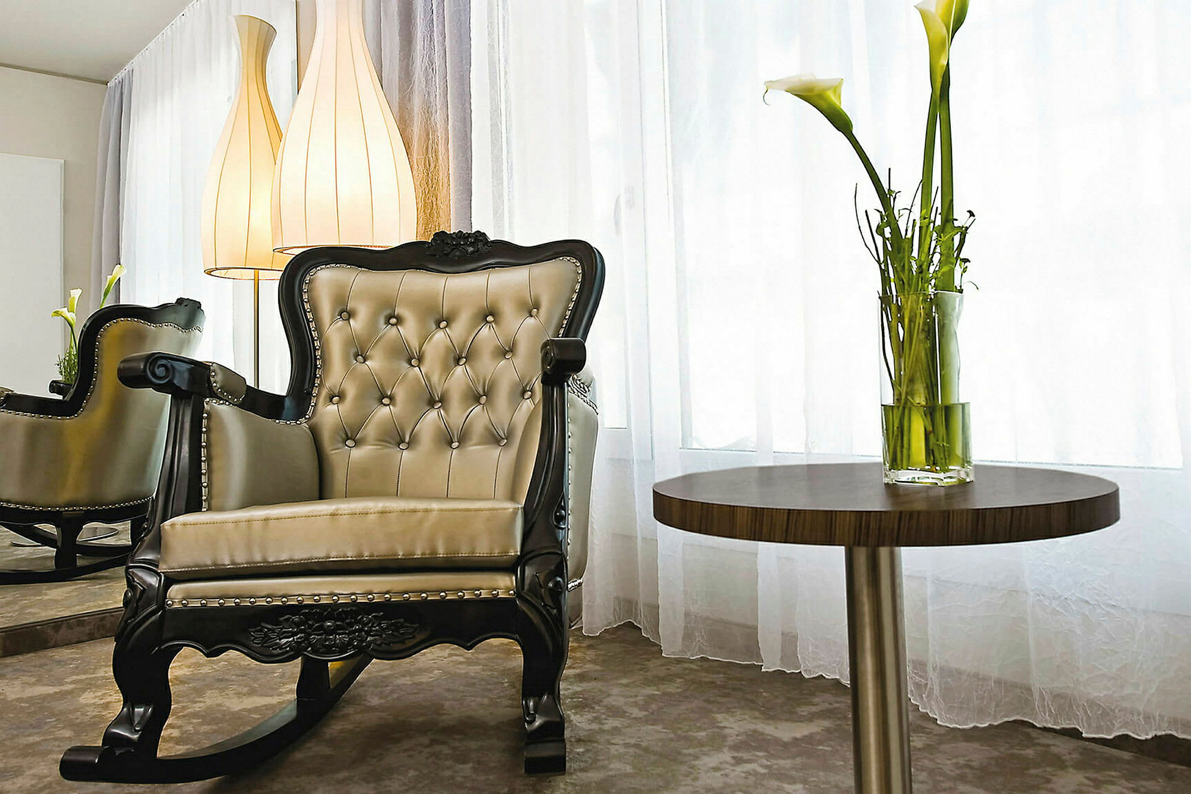 En elegant gyngestol med kapitél og et rundt sidebord med en vase med hvite liljer, i et rom med lyse gardiner. (AI-generert tekst)