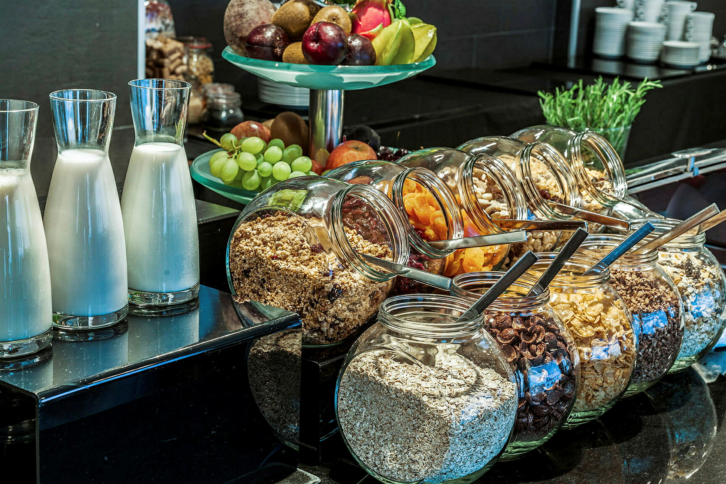 En frokostbuffet med glassflasker med melk, glasskrukker med ulike typer frokostblandinger og müsli, samt en bolle med fersk frukt. (AI-generert tekst)