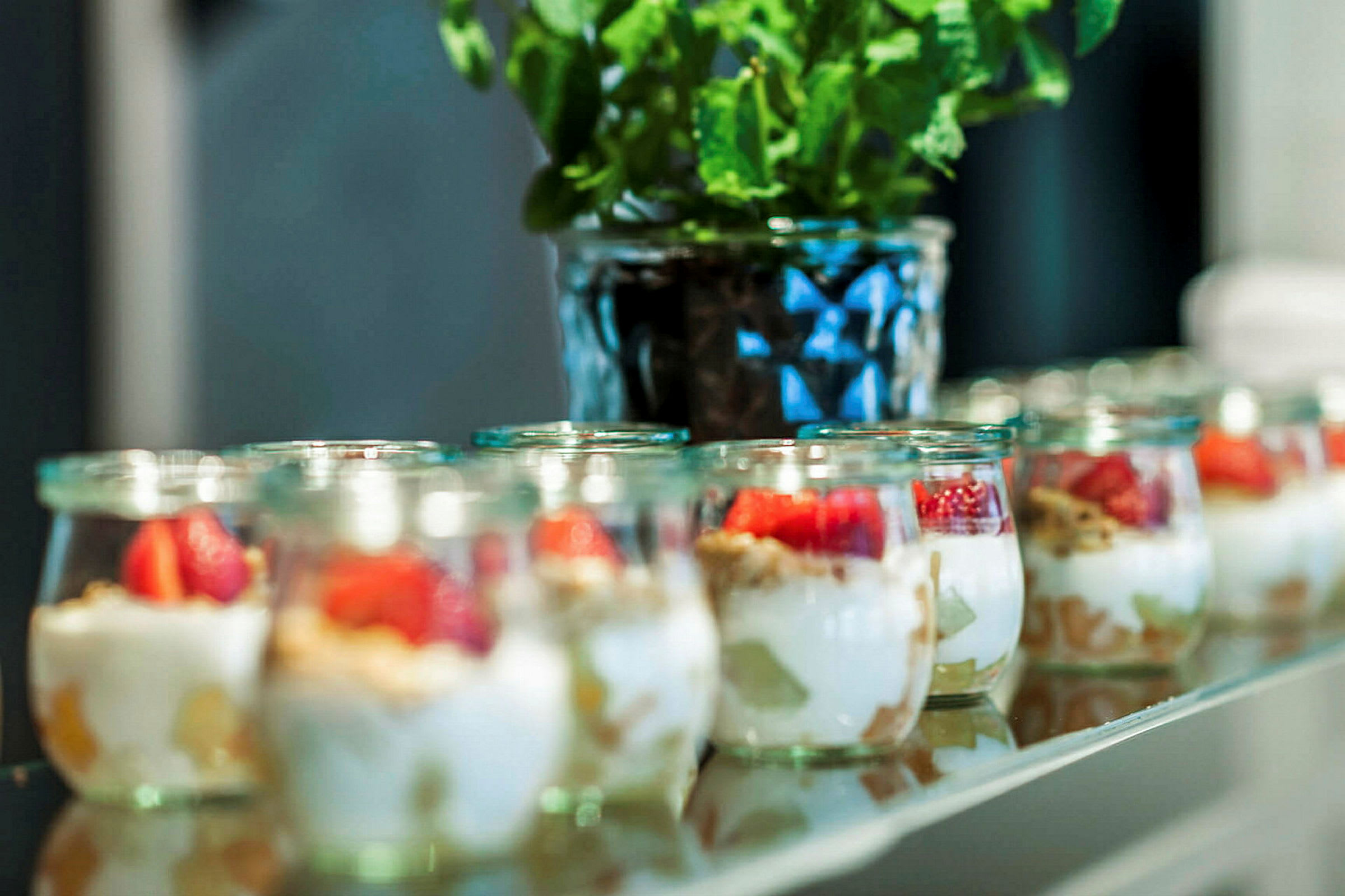 Små glasskrukker med yoghurt, frukt og granola står på rad. I bakgrunnen er en potteplante. (AI-generert tekst)