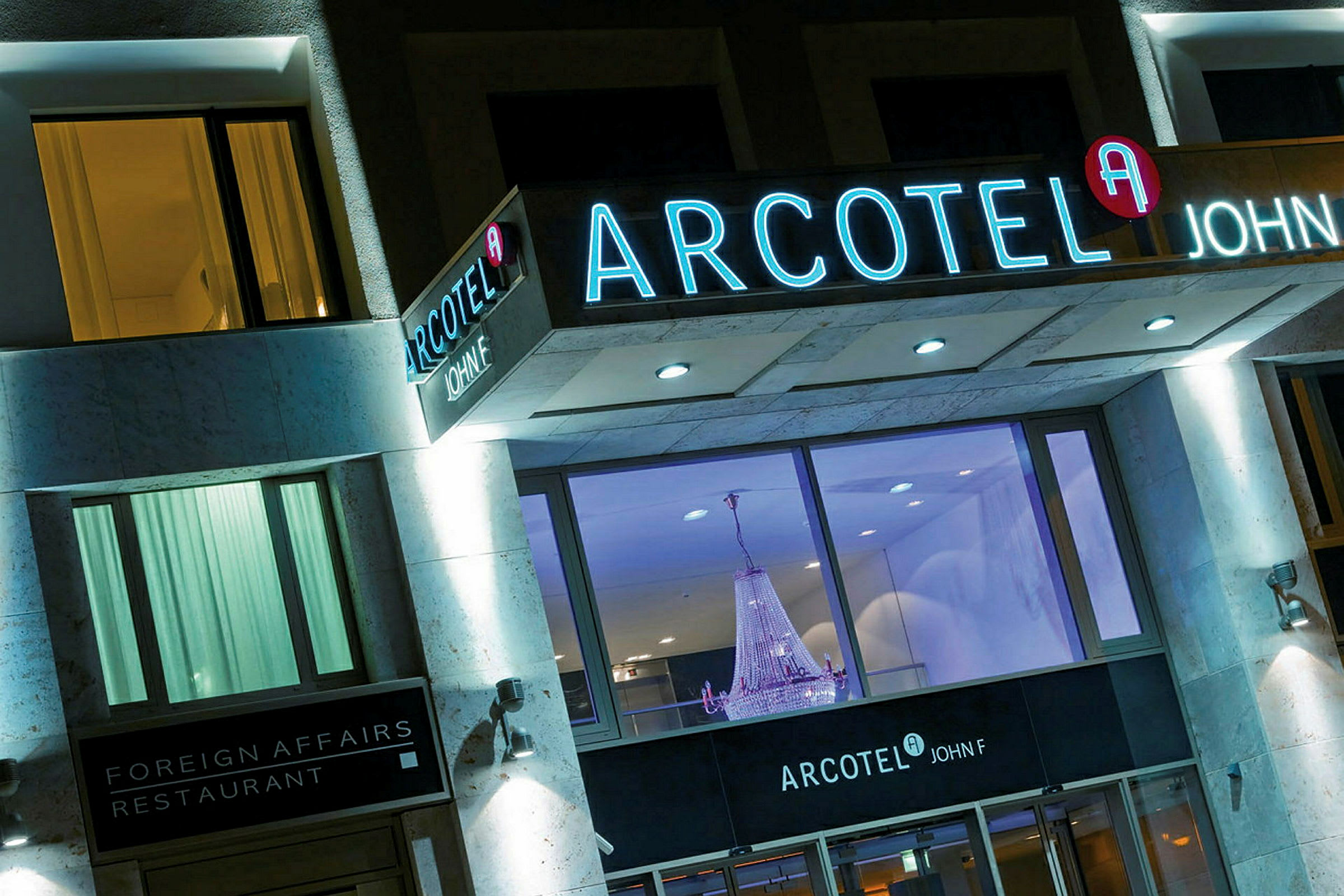 Bilde av en hotellfasade med lysskilt som viser navnet Arcotel John F, samt en restaurant kalt Foreign Affairs. En stor lysekrone er synlig gjennom de store vinduene. (AI-generert tekst)