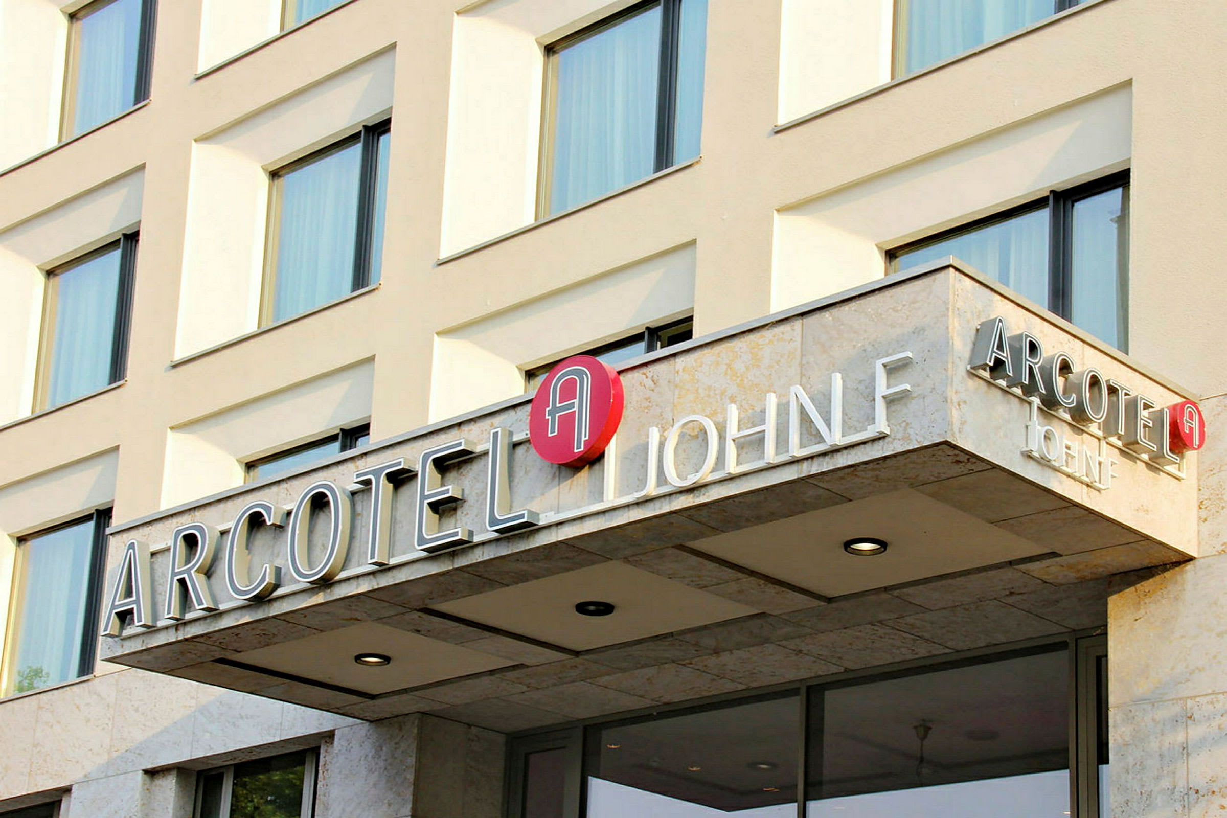 Fasaden til en hotellbygning med skiltet "Arcotel John F". (AI-generert tekst)