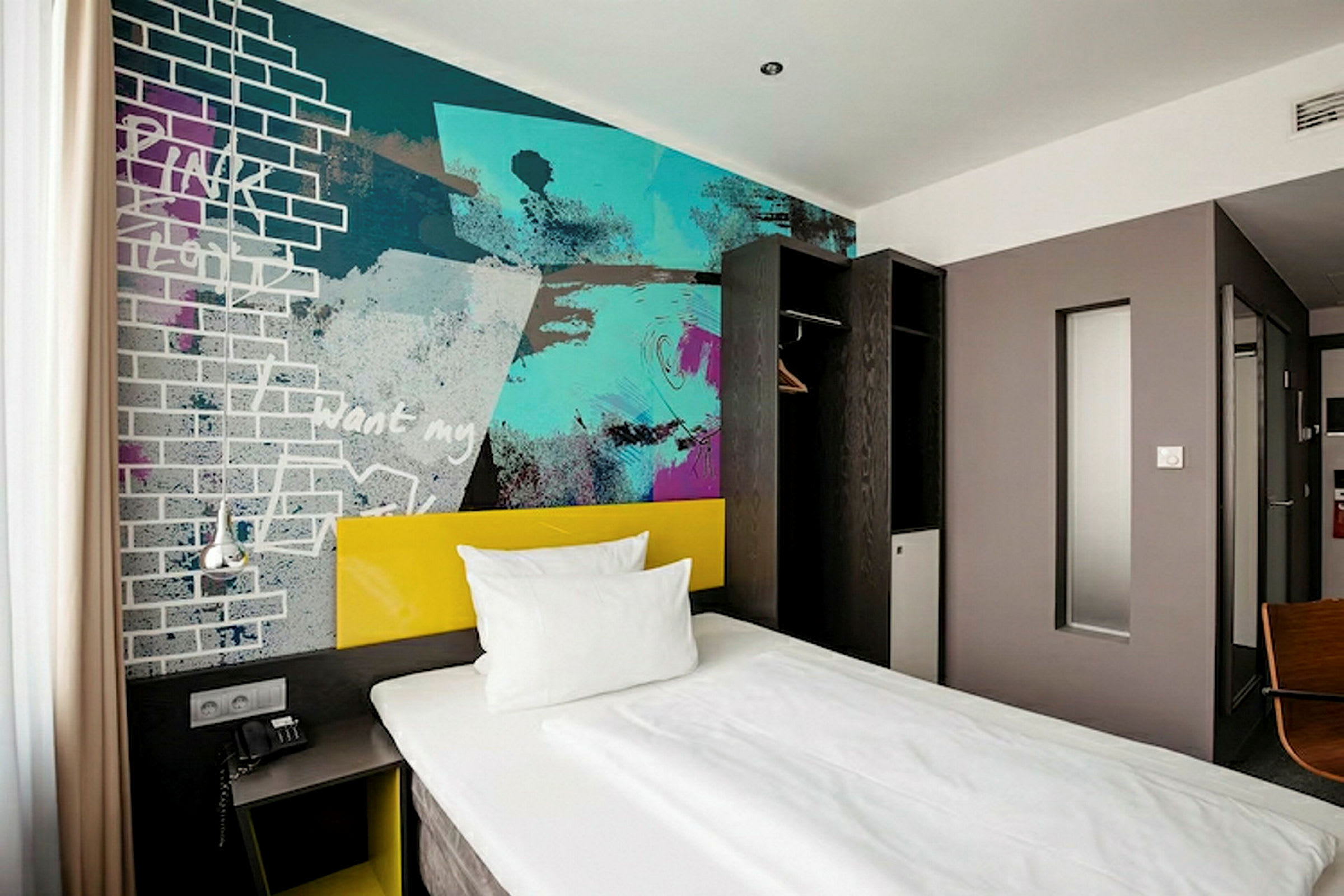 Et hotellrom med en enkeltseng, et fargerikt veggmaleri og minimalistisk interiør. (AI-generert tekst)