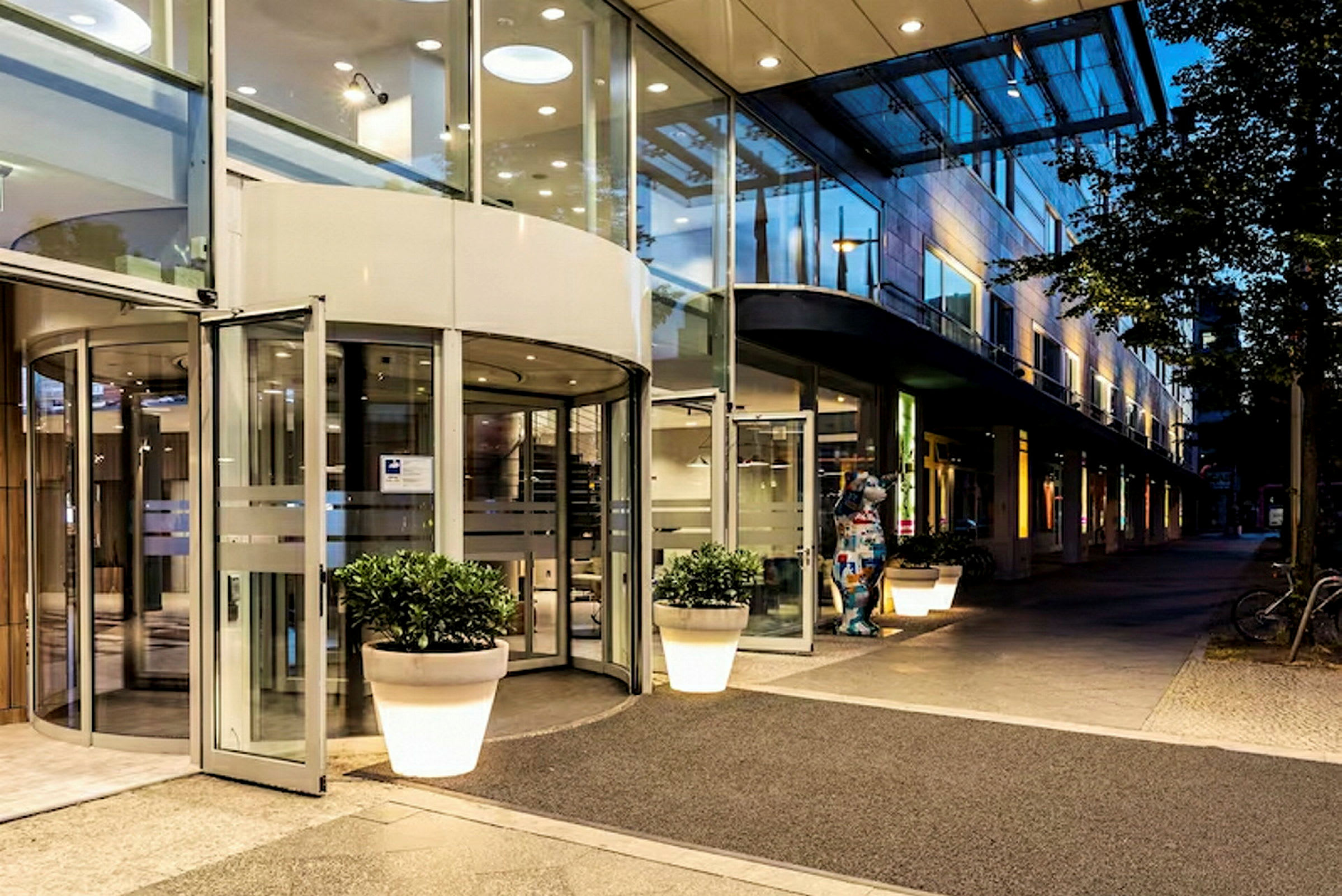 Inngangen til en bygning med glassdører og roterende dør, omgitt av store blomsterkrukker og opplyst under kveldens belysning. (AI-generert tekst)