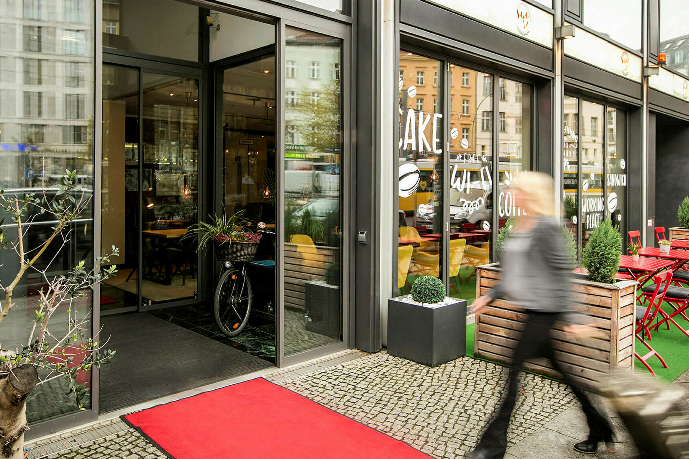 Inngang til en kafé med glassfasade, rød matte på bakken, og en person som passerer utenfor. (AI-generert tekst)