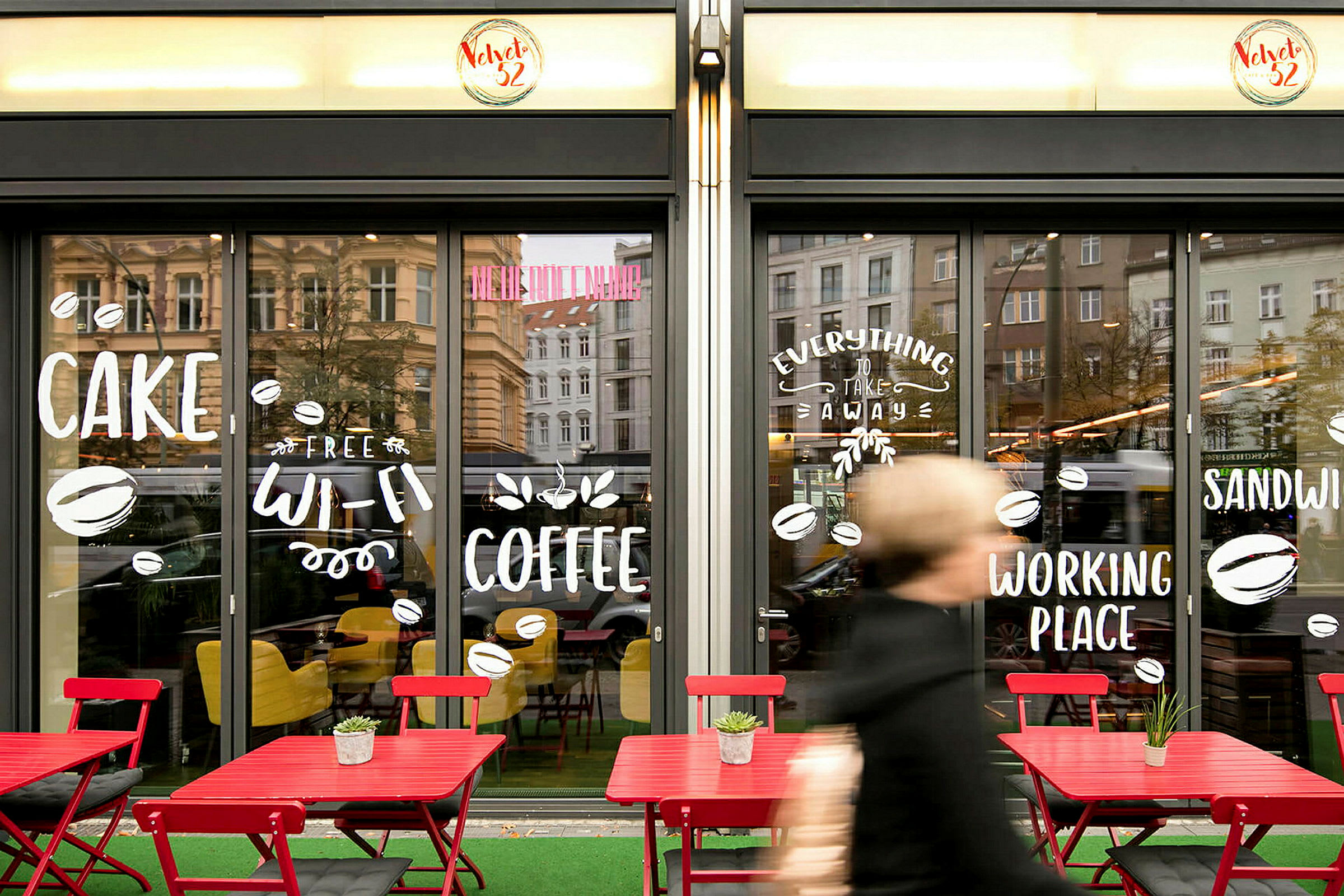 Forsiden av en kafé med store glassvinduer, dekorert med tekst og symboler. Utenfor står røde bord og stoler. (AI-generert tekst)