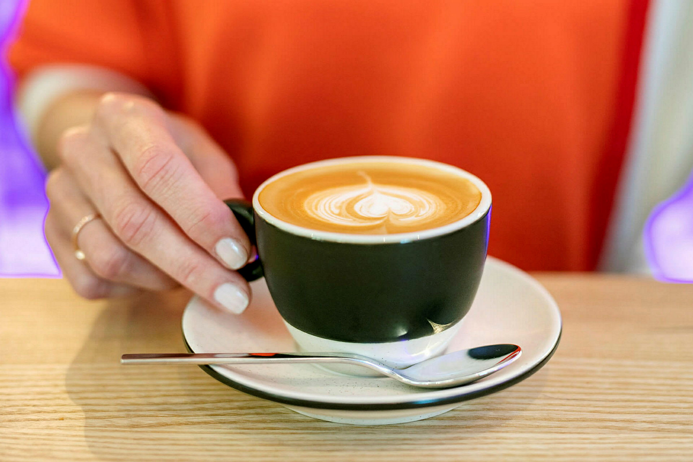 En hånd holder et svart kaffekoppøre med en skål og en skje på et trebord. Cappuccino med lattekunst på toppen. (AI-generert tekst)