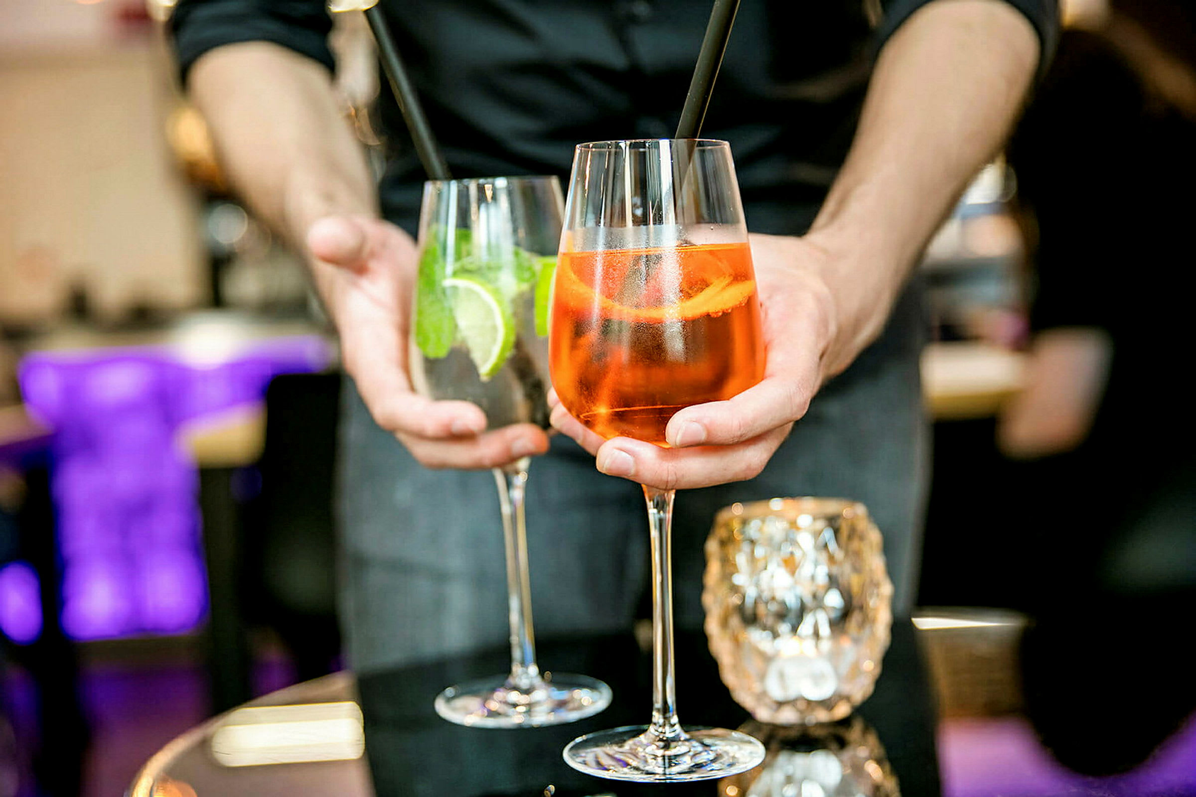 To drinker holdes frem av en person, et glass inneholder en oransje cocktail og det andre en klar cocktail med lime og mynte. (AI-generert tekst)