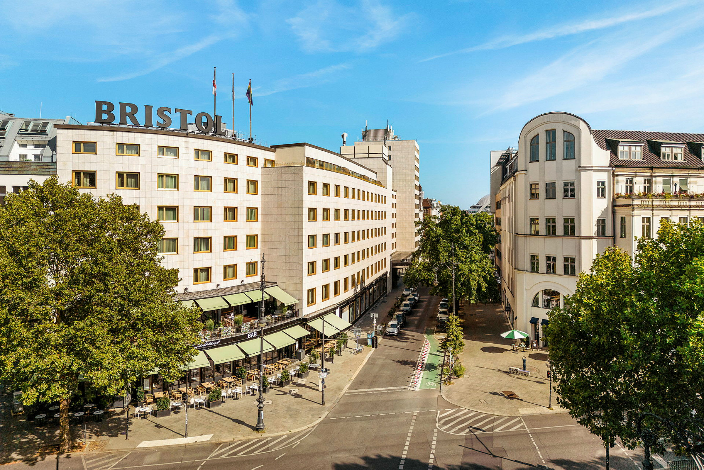 Et bylandskap som viser en bygning med teksten "Bristol" på taket, omgitt av trær og en gate med uteservering. (AI-generert tekst)