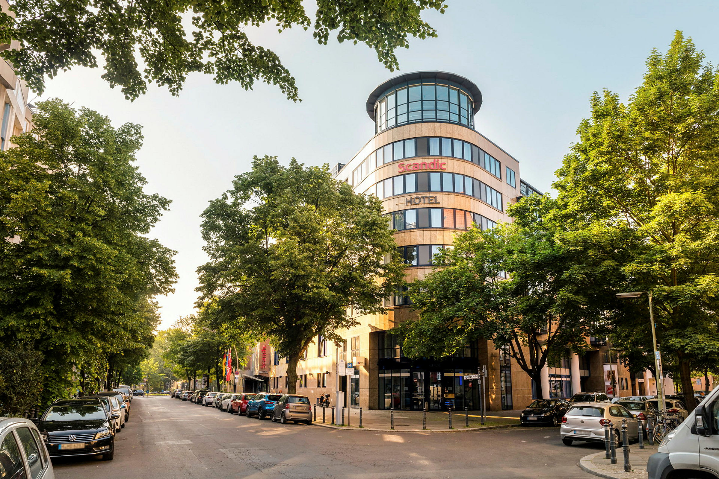 En byutsikt med fasaden til et Scandic-hotell omgitt av trær og parkerte biler. (AI-generert tekst)