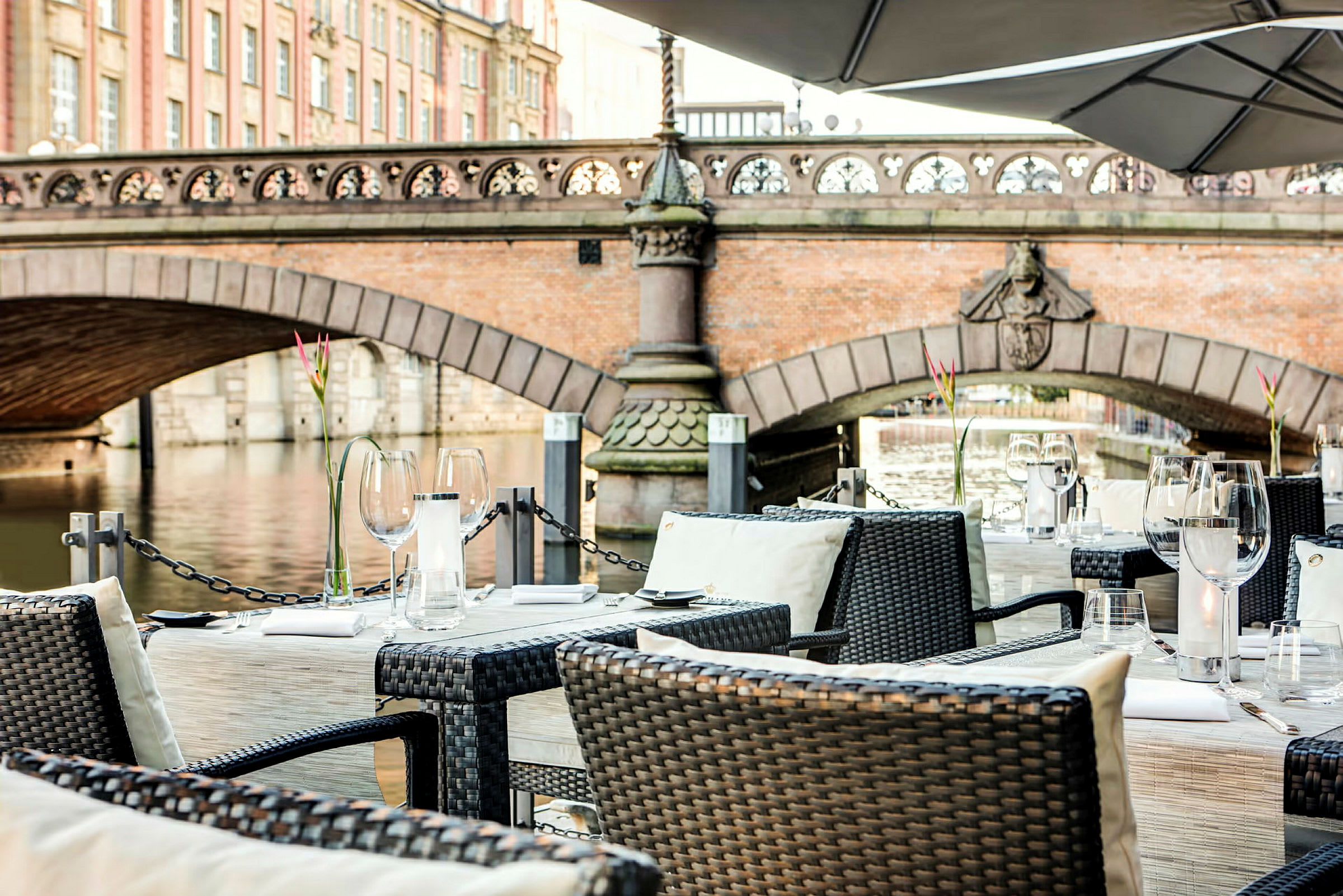 Pent oppsatte bord på en terrasse med utsikt over en bro og en elv. (AI-generert tekst)