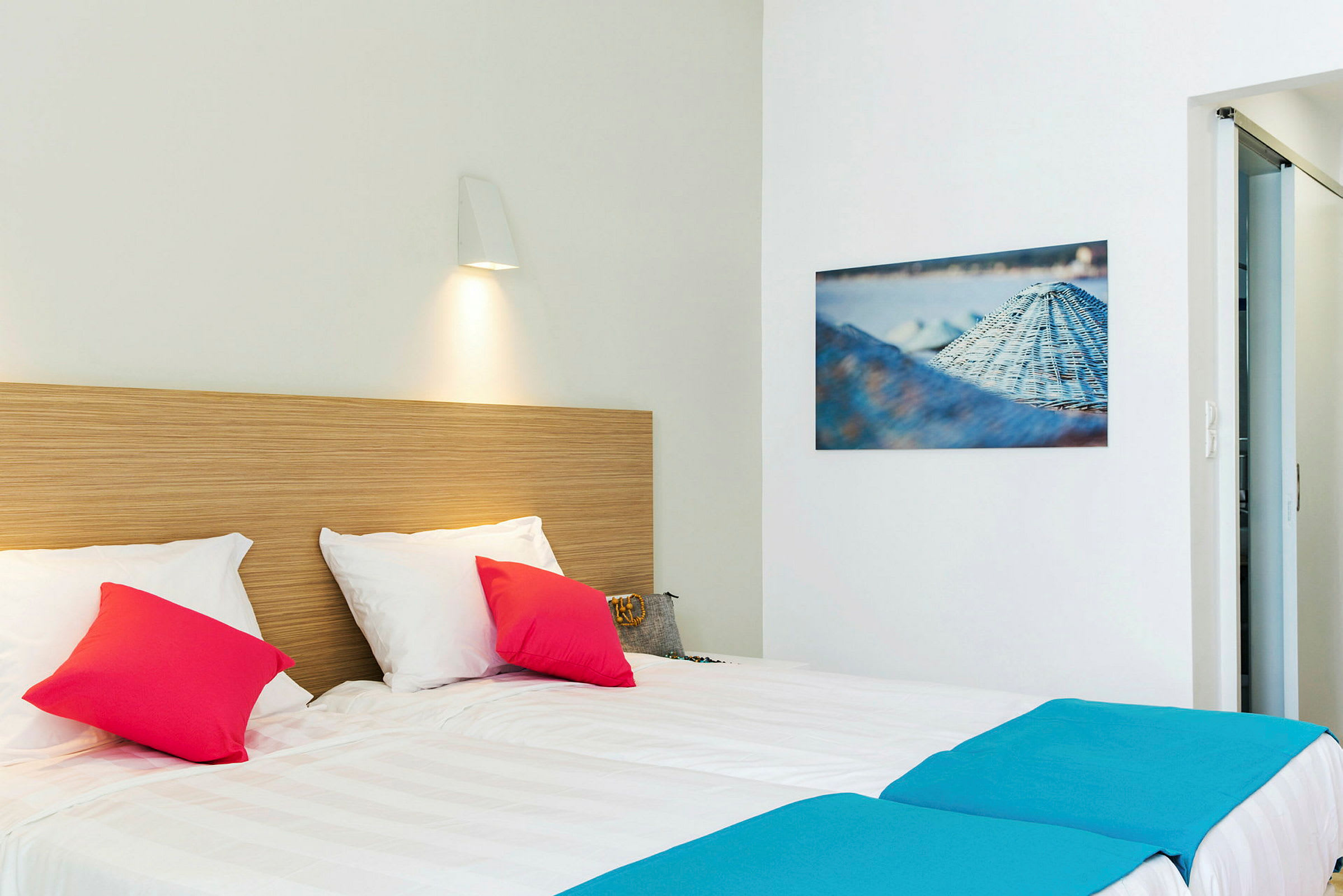 Hotellrom med dobbeltseng oppredd med hvitt sengetøy, dekorative fargerike puter og en hodegavl av tre. På veggen henger et maleri og en lampe lyser ned mot sengen. (AI-generert tekst)
