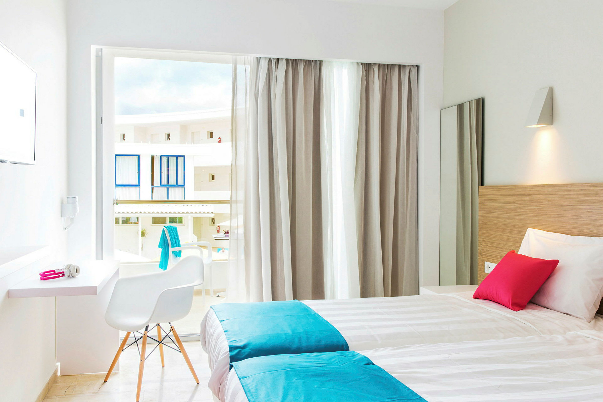 Et lyst hotellrom med to enkeltsenger, hvite og turkise sengetøy, en rosa pute. Vinduet har lyse gardiner og åpner mot balkonger. (AI-generert tekst)
