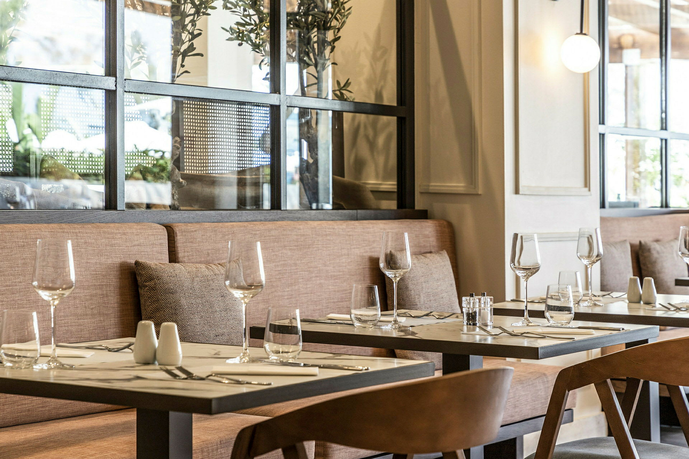 Et restaurantbord med glass og bestikk på en duk, omgitt av stoler og en sofa med puter. (AI-generert tekst)