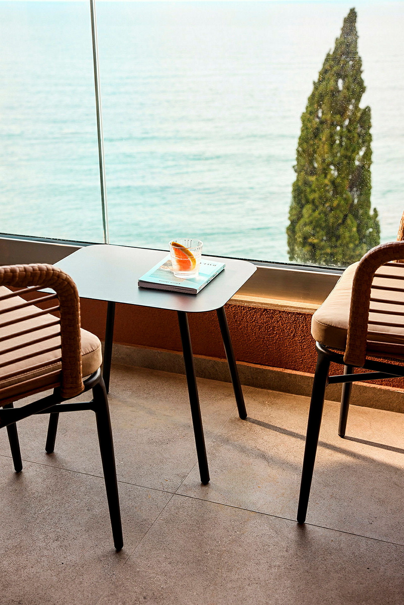 To stoler og et bord med et glass og en bok står på en terrasse med utsikt over havet og et tre. (AI-generert tekst)