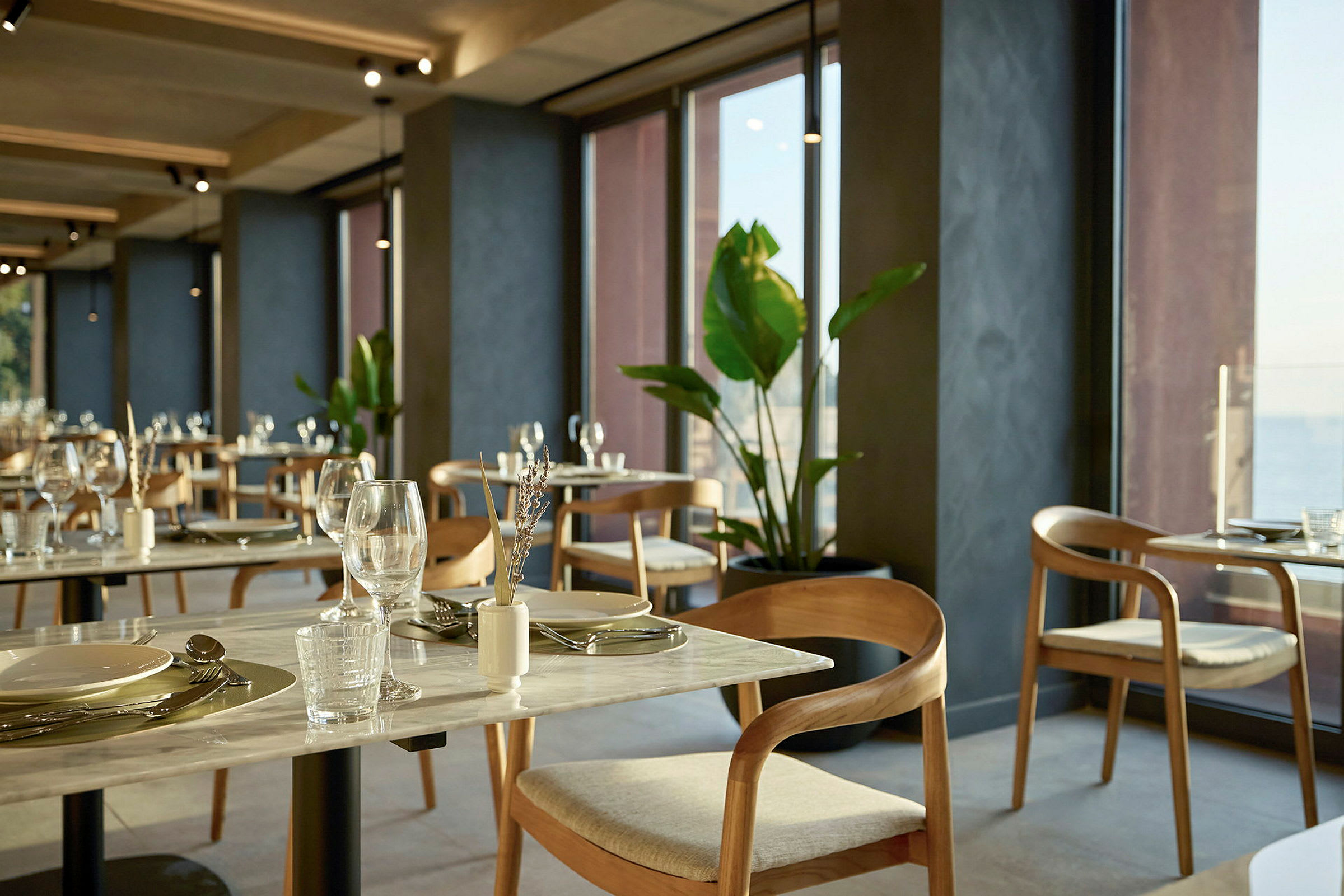 En restaurant med bord og stoler ved store vinduer, dekorert med planter. (AI-generert tekst)