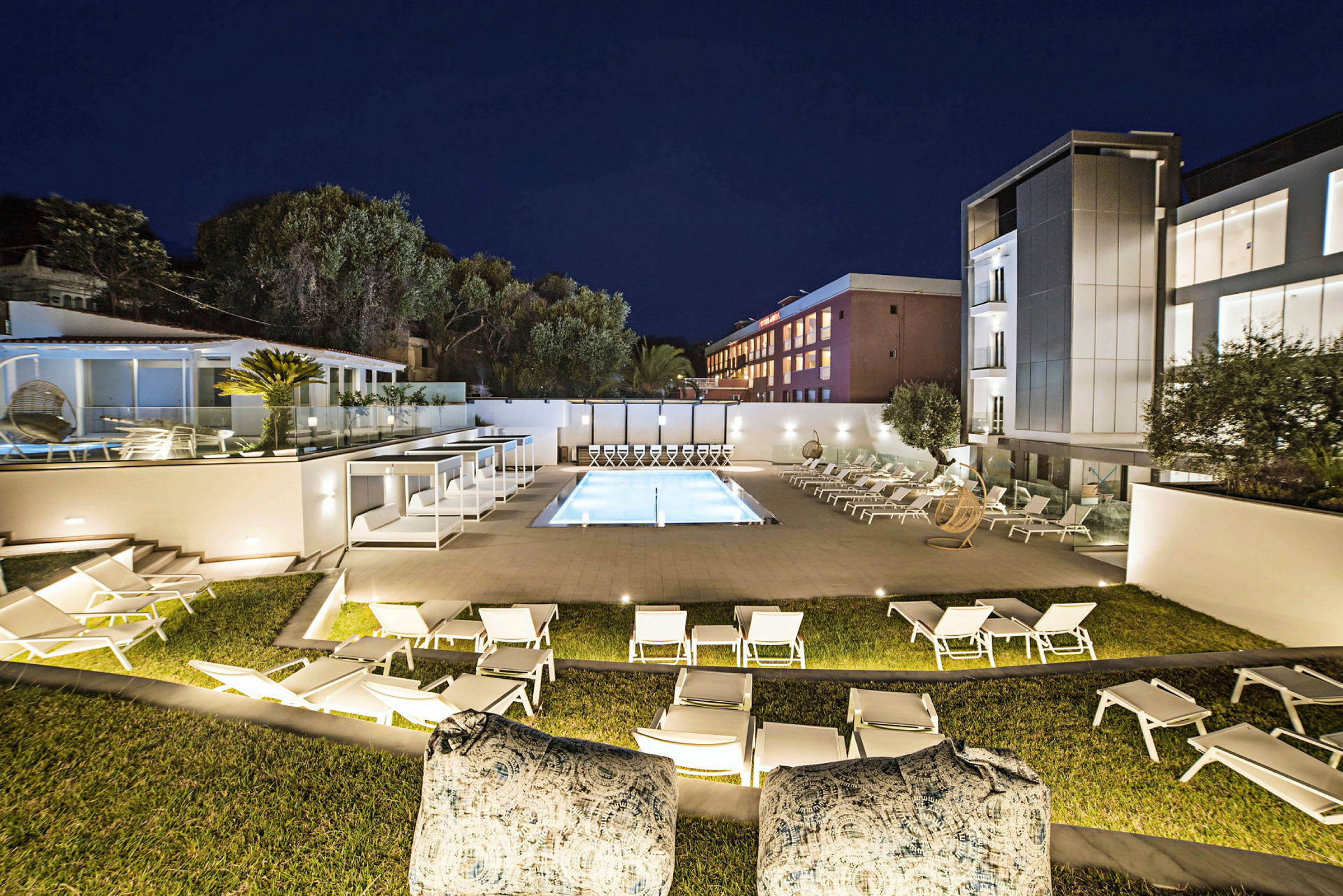 Et opplyst hotellområde på kvelden, med et basseng omgitt av solsenger og moderne bygninger i bakgrunnen. (AI-generert tekst)