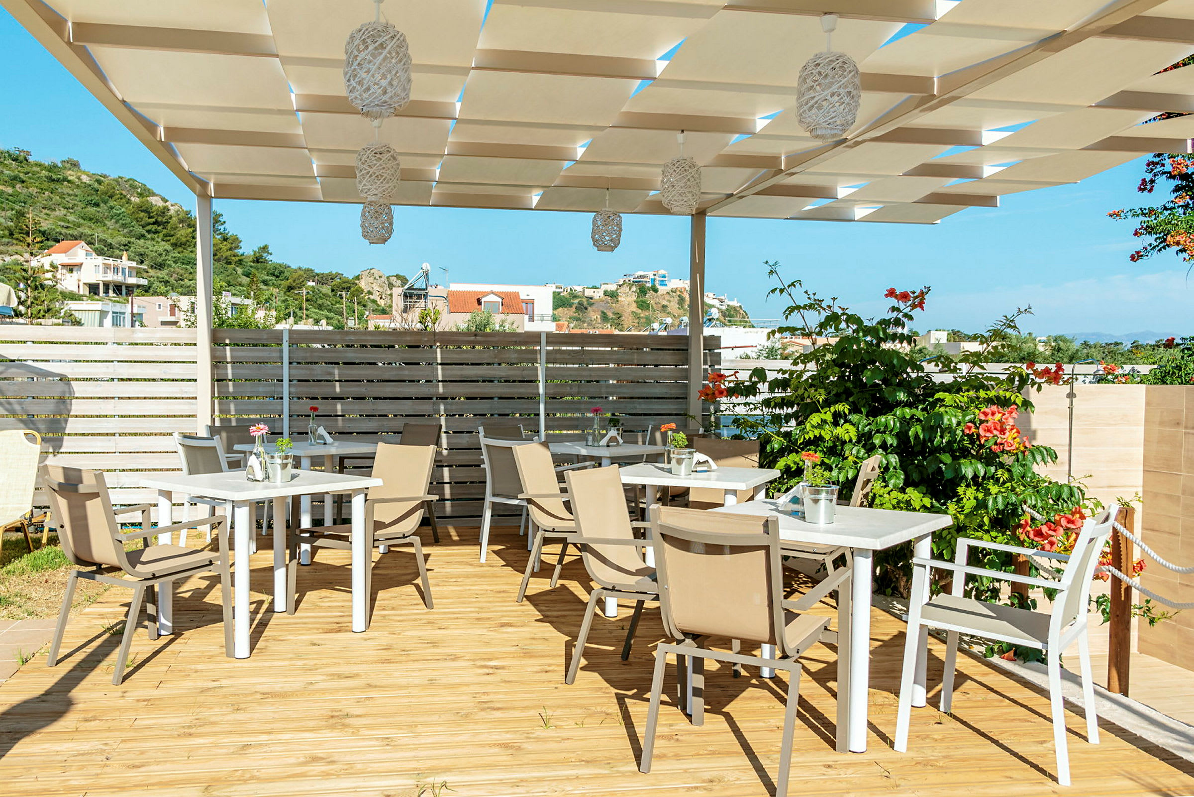 En terrasse med tremøbler og tak bestående av hvite paneler, omgitt av planter og med utsikt over et landlig område. (AI-generert tekst)