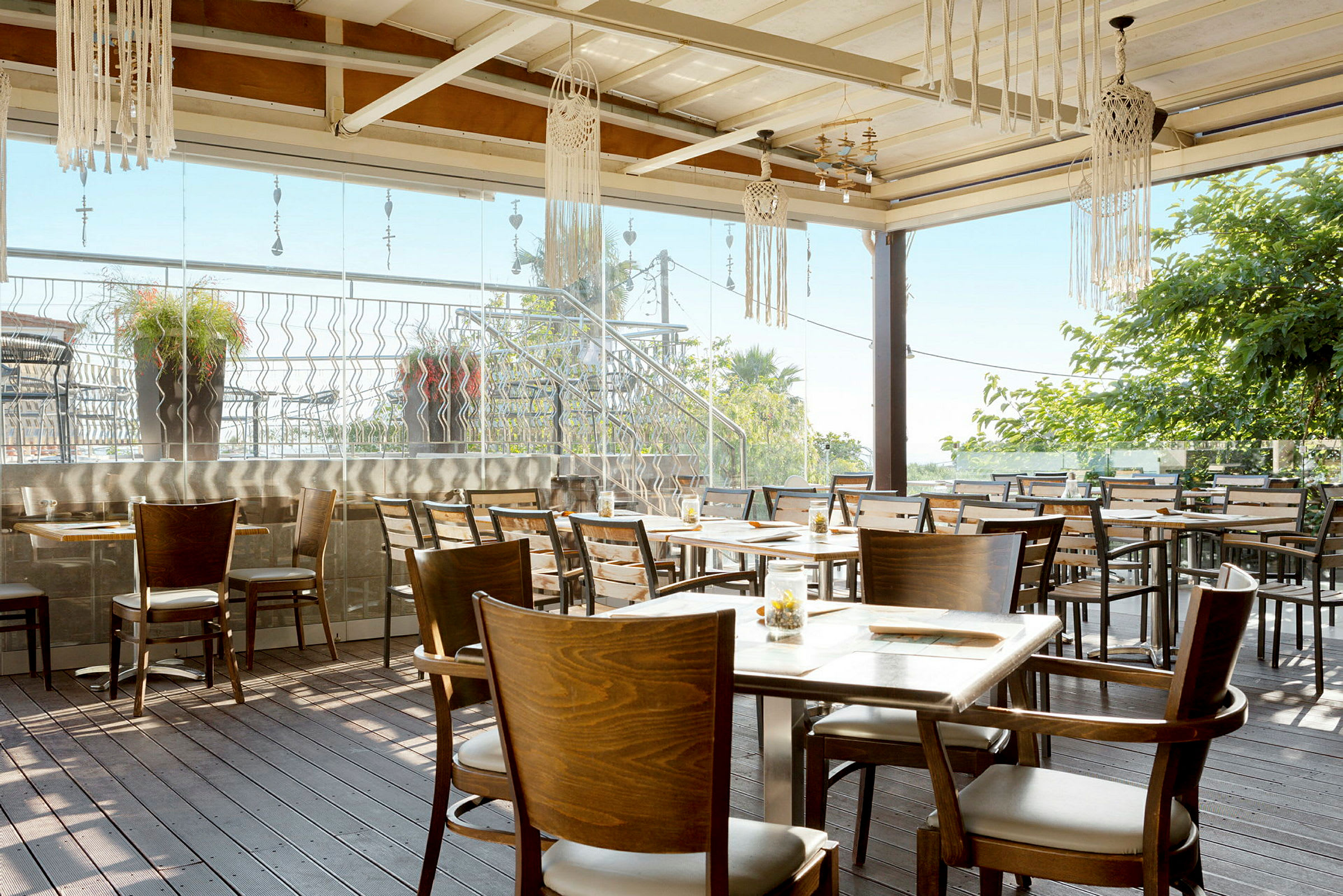 Bilde av en utendørs terrasse med trebord og stoler. Dekorasjoner henger fra taket, og det er grøntområder i bakgrunnen. (AI-generert tekst)