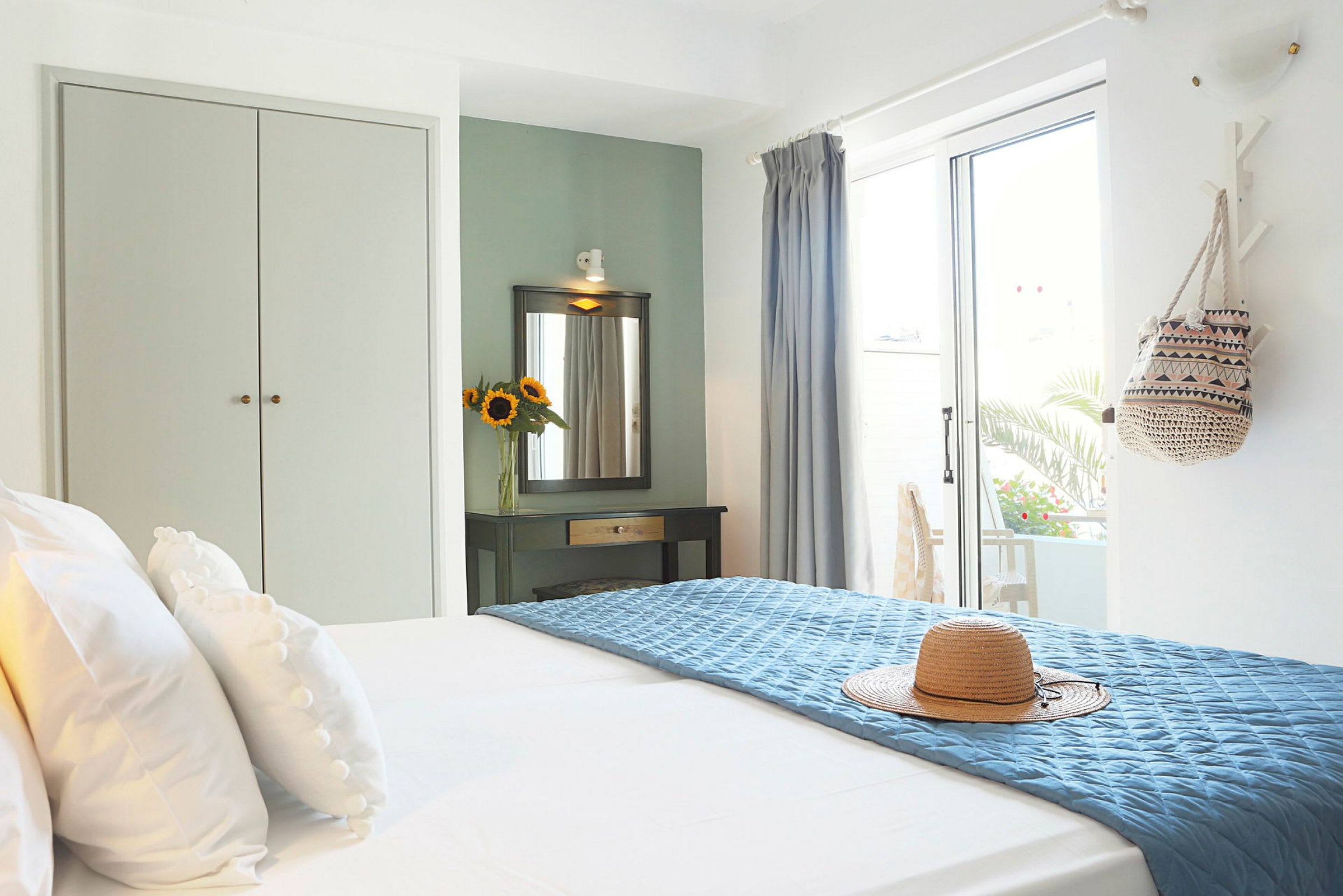 Et lyst hotellrom med en oppredd seng, en solhatt på blått sengeteppe og et dekorasjonsbord med speil og en vase med solsikker. (AI-generert tekst)