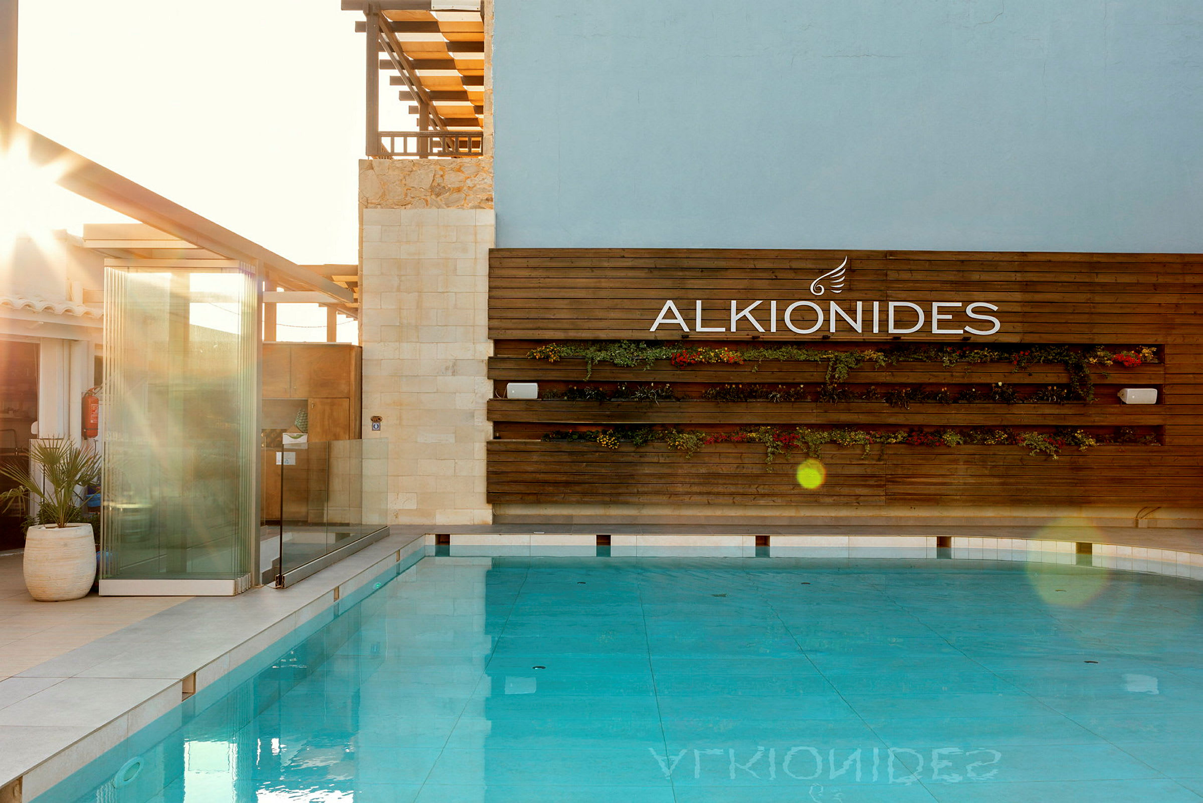 Bilde av et basseng med en trevegg i bakgrunnen der det står "ALKIONIDES" og dekorasjoner med planter. (AI-generert tekst)