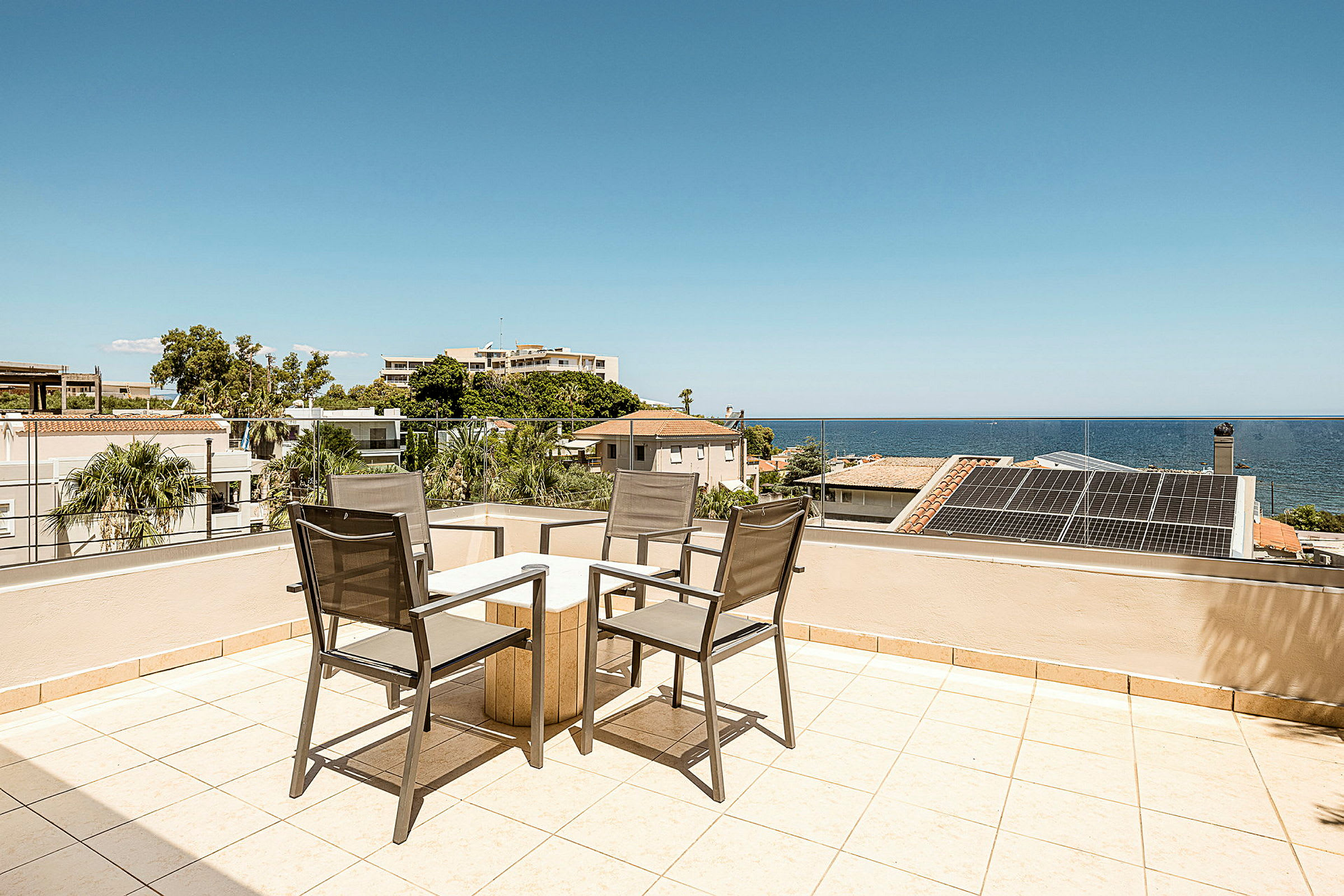 Utsikt over en terrasse med bord og stoler, havet og bygninger i bakgrunnen. (AI-generert tekst)