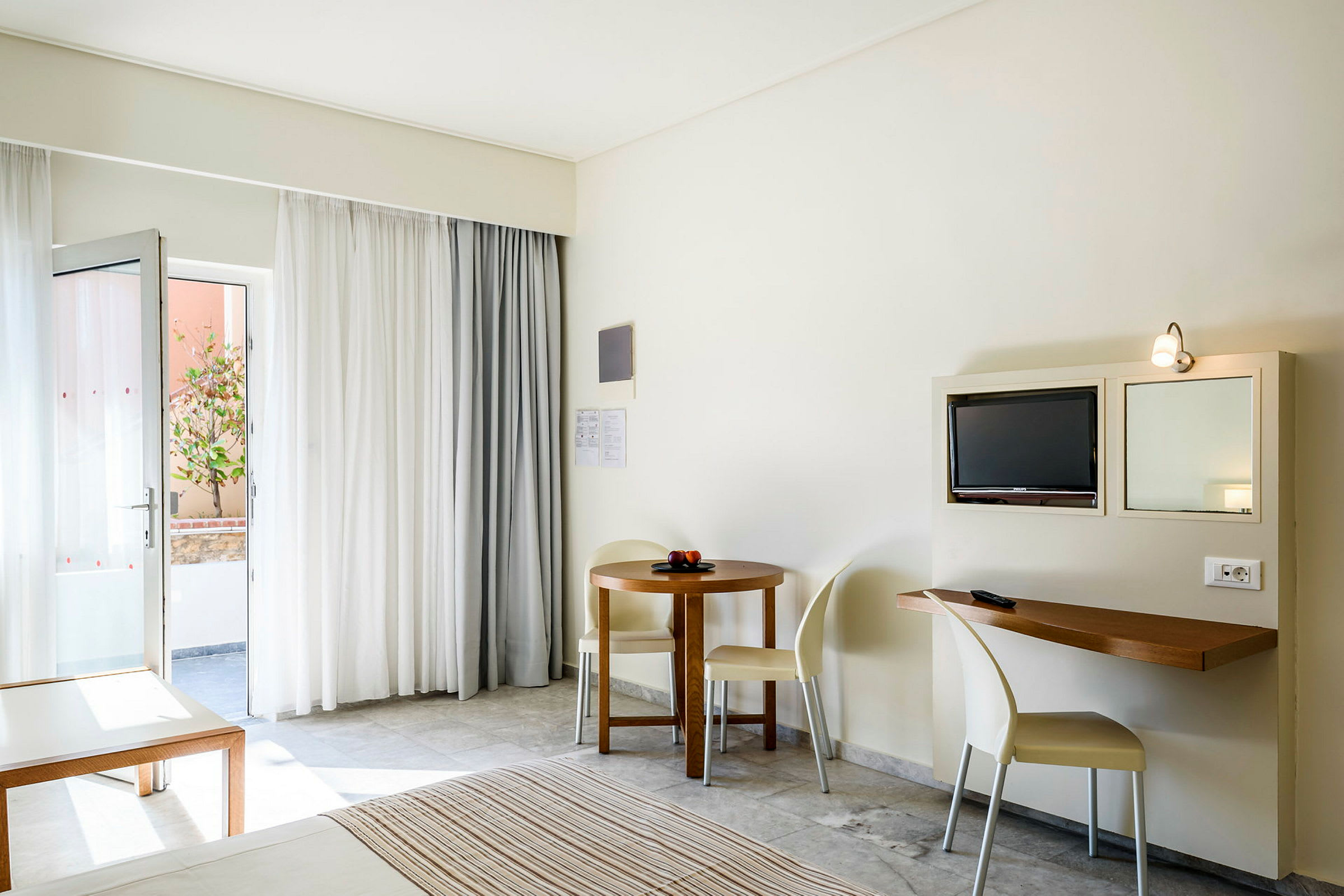 Et hotellrom med en åpen dør, en liten spiseplass og en TV montert på veggen. (AI-generert tekst)