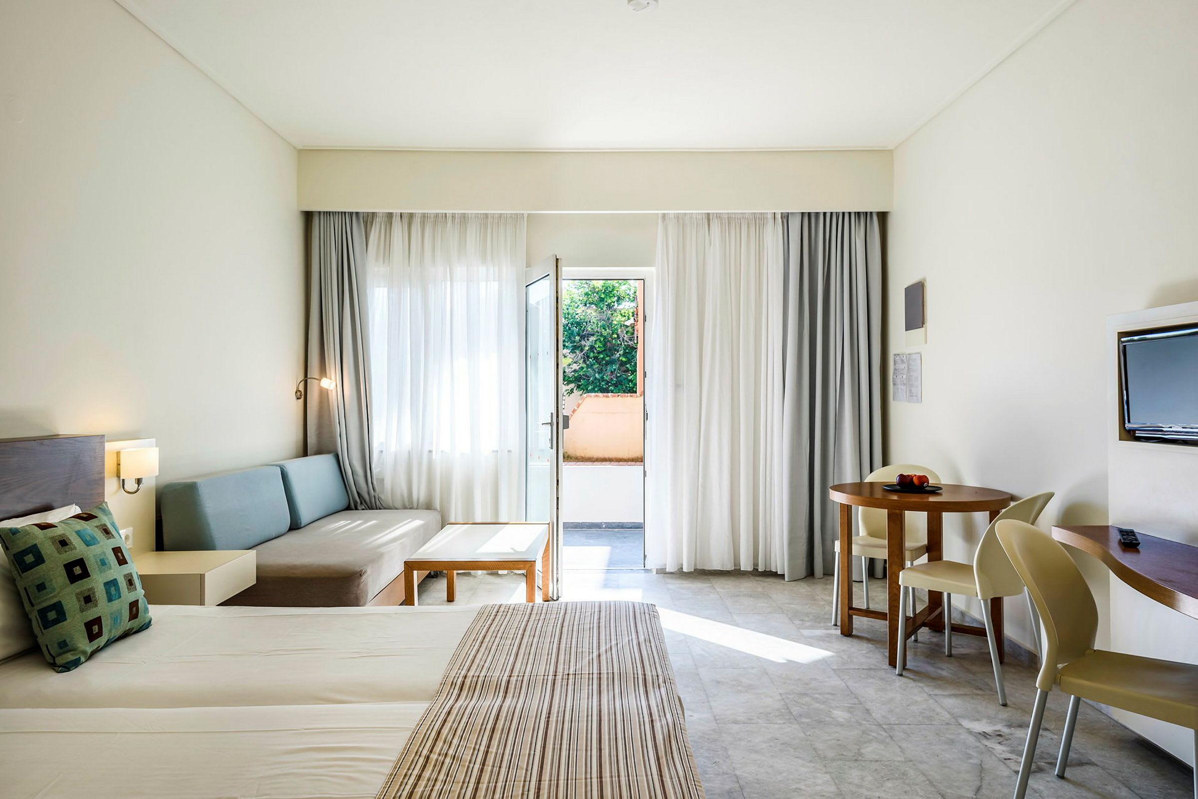 Hotelrom med oppredd seng, sittegruppe, rundt spisebord og balkongdør. (AI-generert tekst)