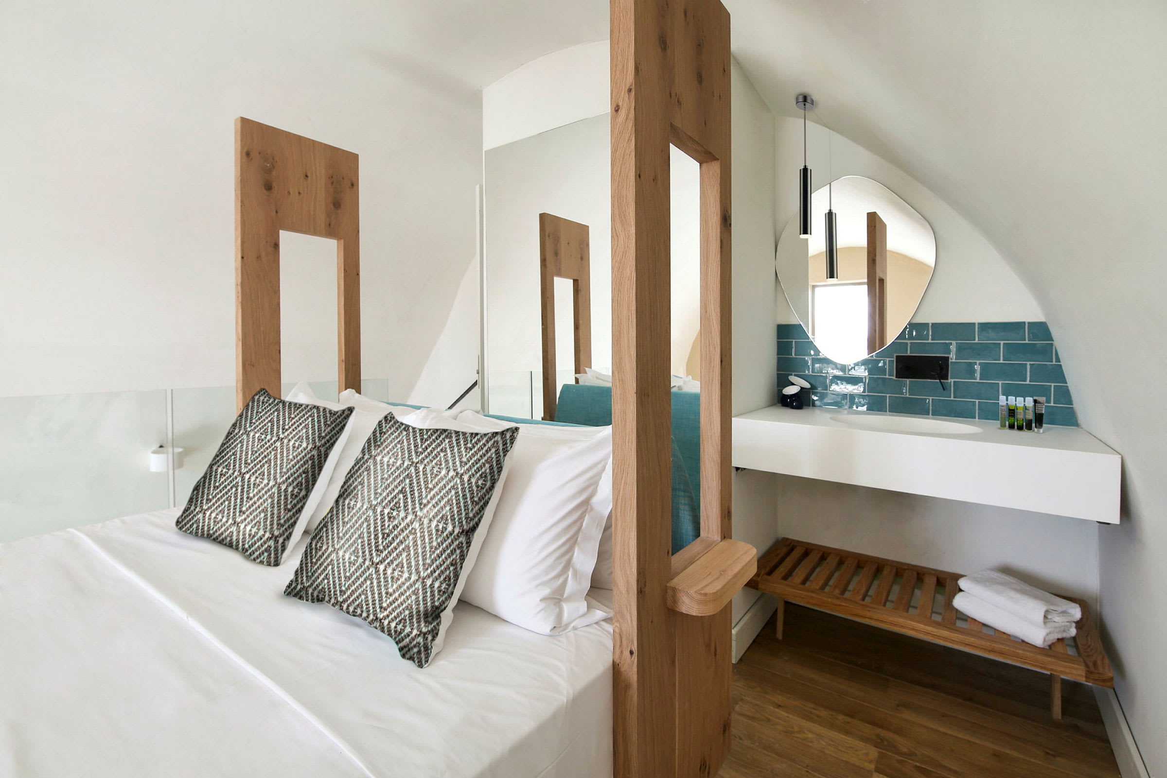 Et hotellrom med en stor seng, to pynteputer og et moderne baderomsområde med et rundt speil og en trebenskrakk under en hvit hylle. (AI-generert tekst)