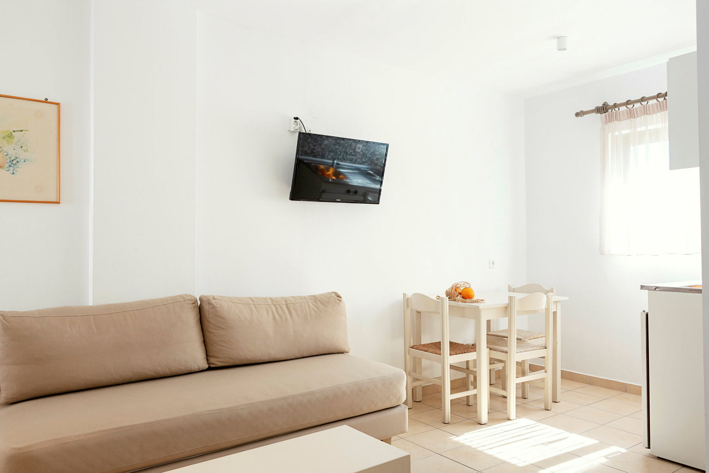 Et lyst rom med beige sofa, veggmontert TV, hvitt spisebord med stoler og et vindu med lyse gardiner. (AI-generert tekst)