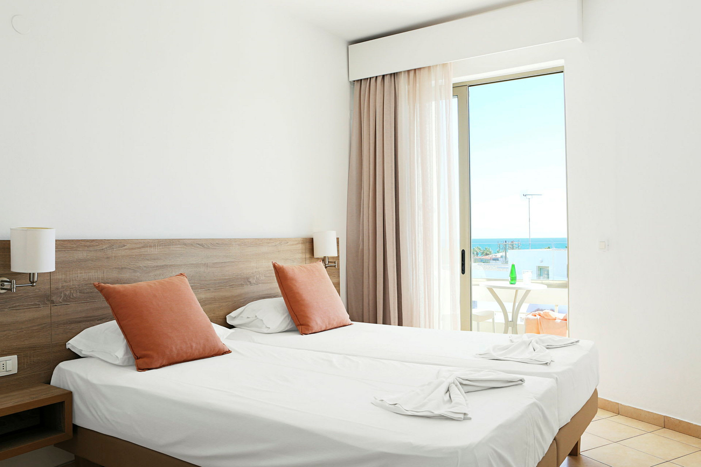 Hotellrom med to enkeltsenger, oransje puter og en balkong med havutsikt. (AI-generert tekst)