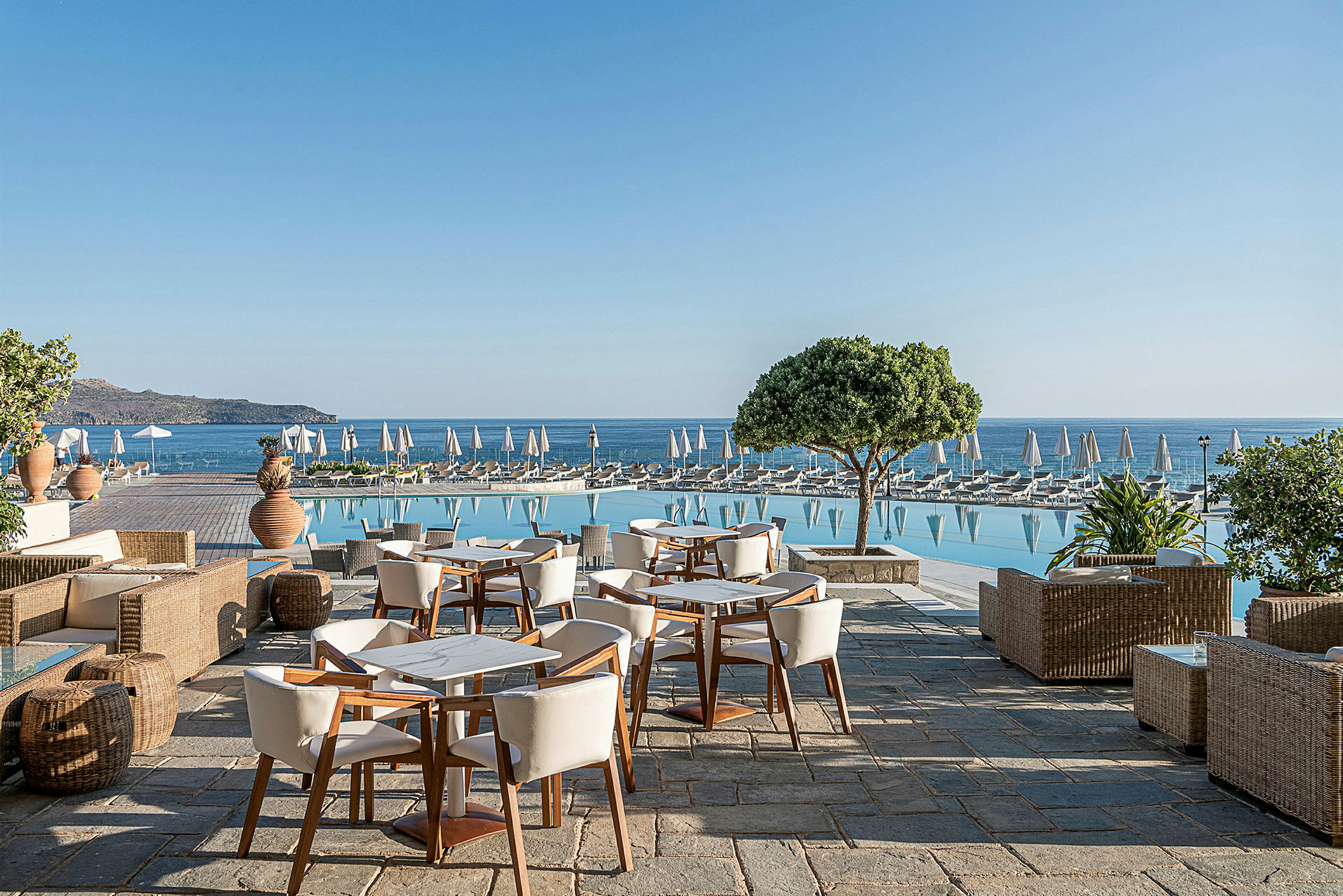 Utendørs terrasse med bord og stoler, et basseng med solsenger ved siden av, og havet i bakgrunnen. (AI-generert tekst)