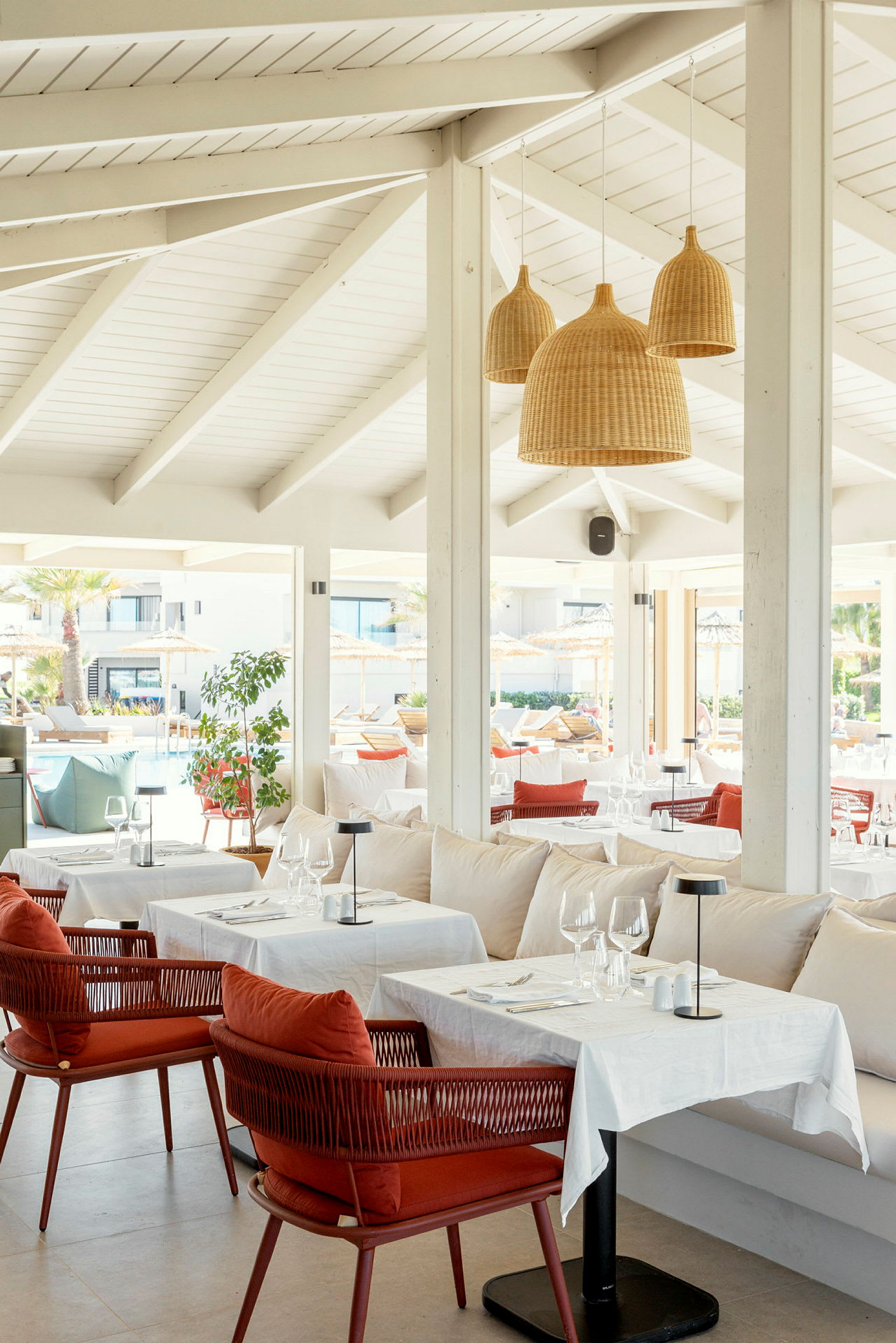 Interiør av en restaurant med hvite takbjelker, hengende rottinglamper samt dekkede bord og stoler. (AI-generert tekst)