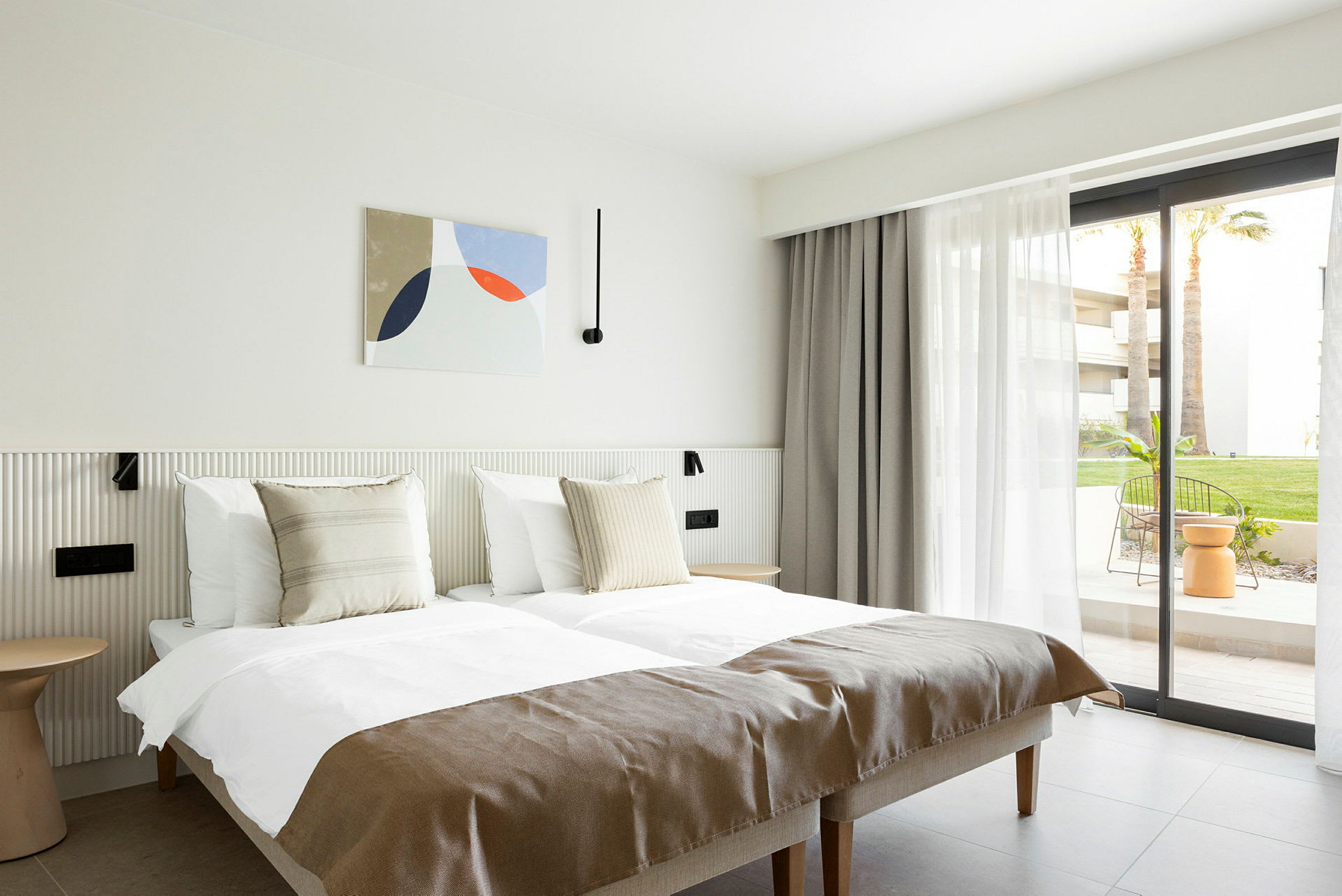 Et lyst hotellrom med to enkeltsenger, nøytrale detaljer og en vindusdør mot en uteplass. (AI-generert tekst)